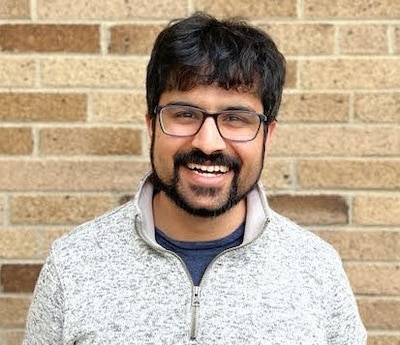 Headshot of a man with a dark beard wearing glasses. He’s wearing a heather gray sweater. 