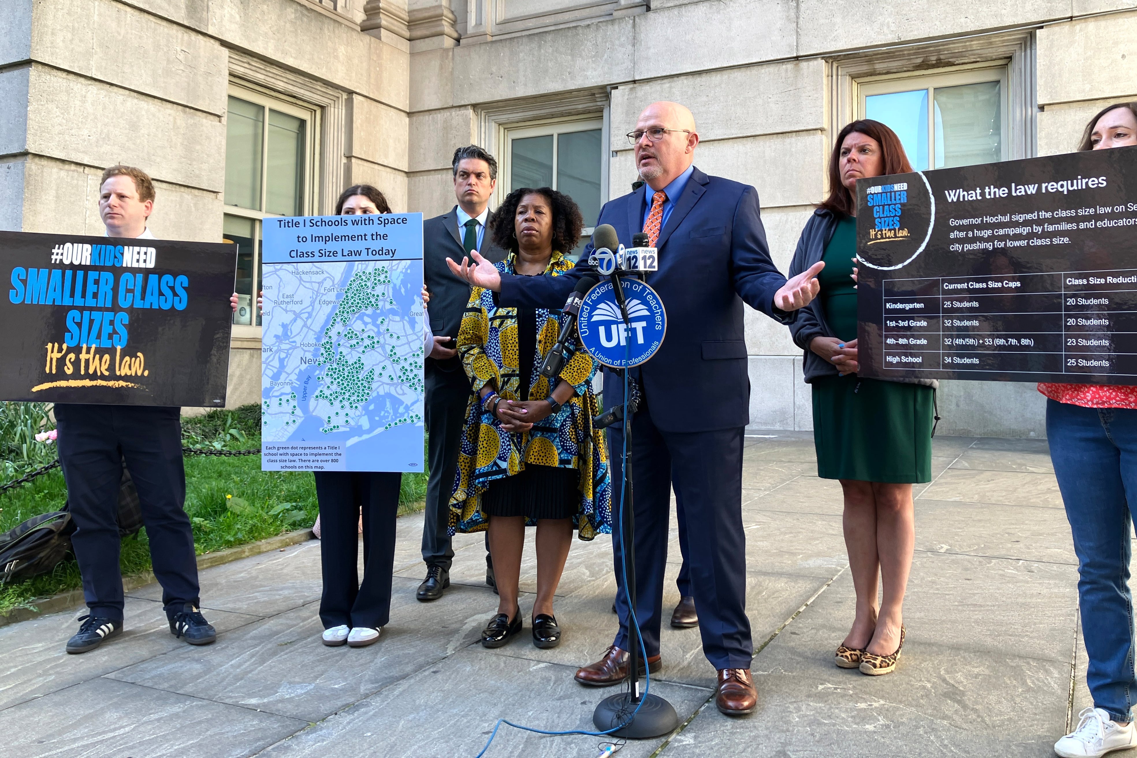 A man in a blue suit gestures in front of a microphone surrounded by other people with signs.