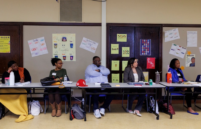 Five people sit in a row at a long table with colorful posters and signs on the wall in the background.