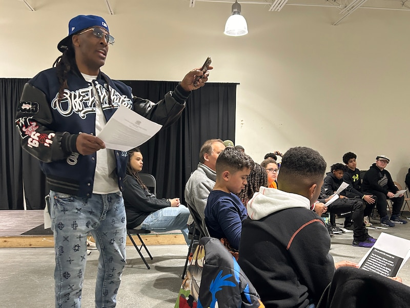 An adult man wearing a ball cap and jacket speaks to a circle of students in a room with a black curtain in the background.