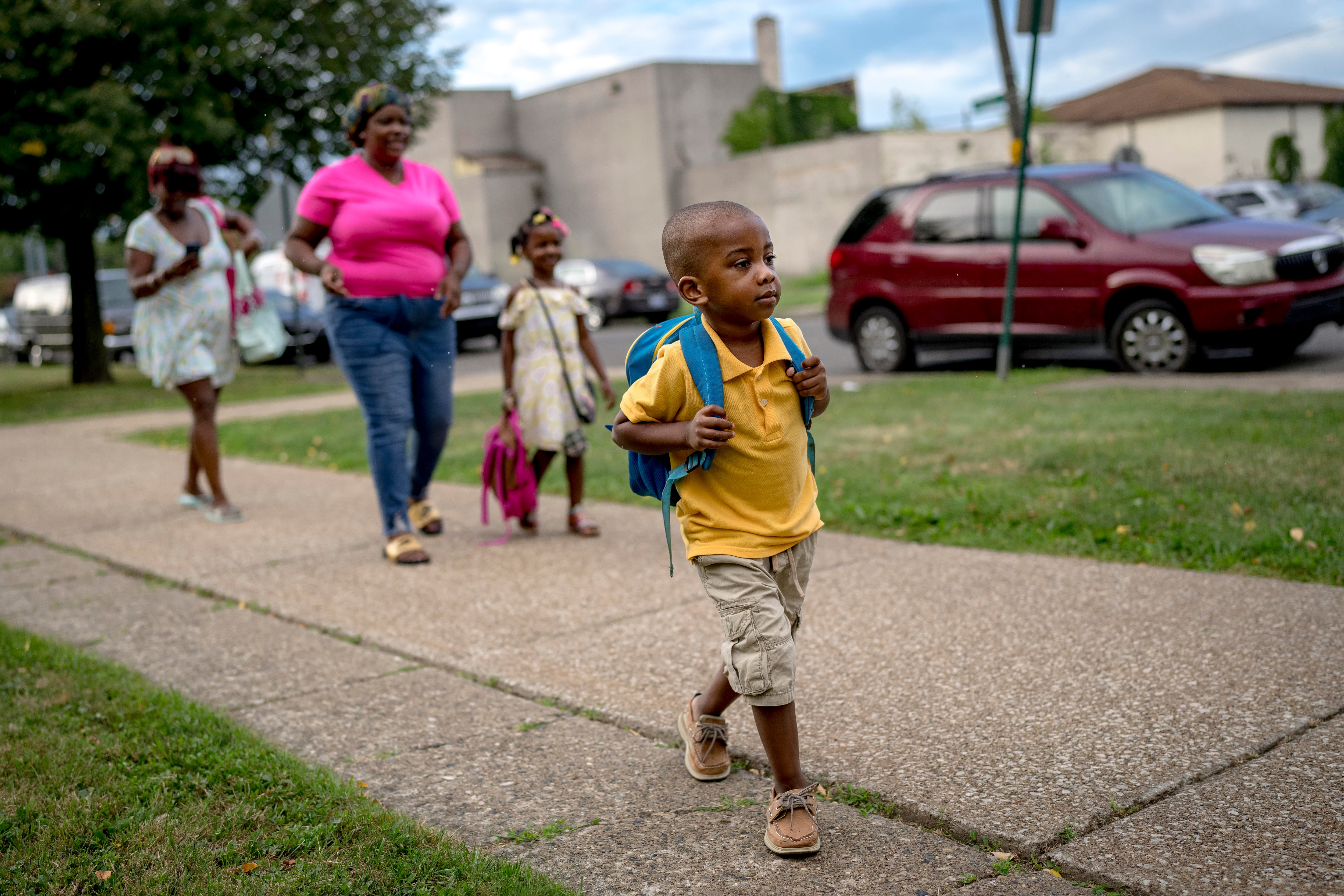 A child wearing a backpack walks toward a school building. Other children and parents can be seen behind him.