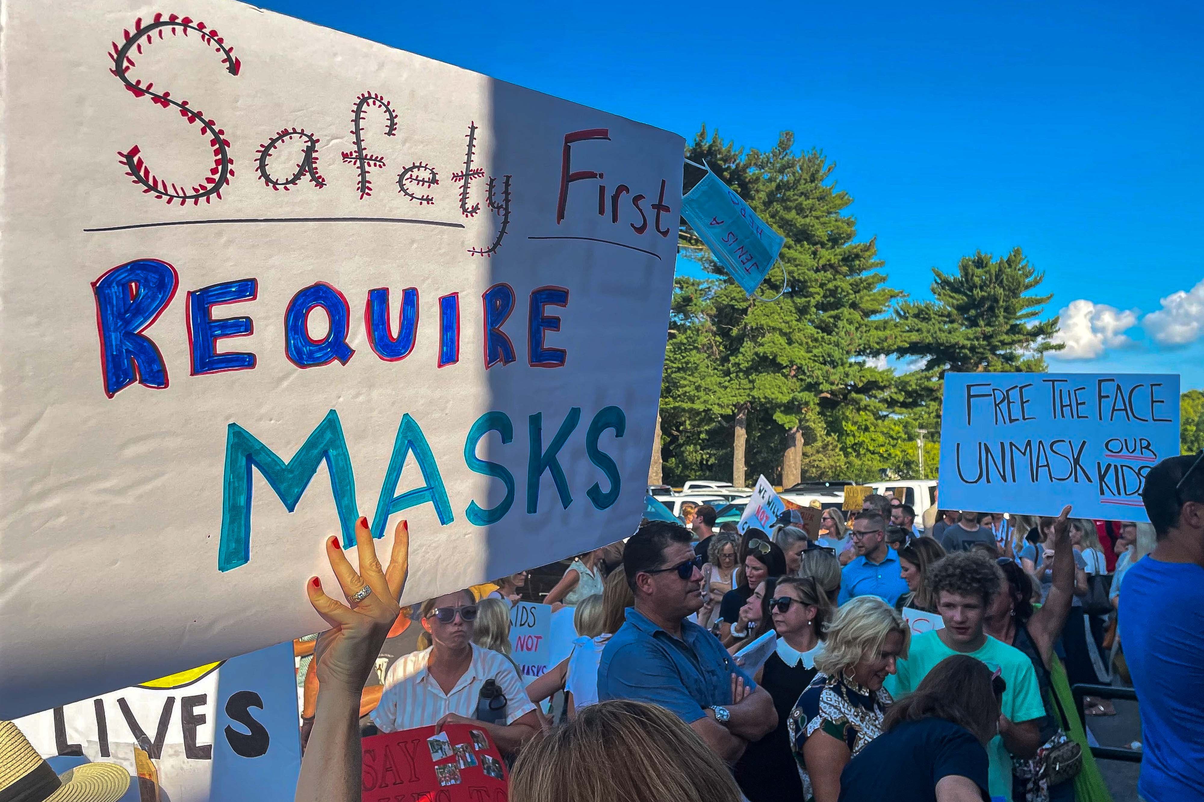 A crowd of people hold up signs for and against school mask mandates during a daytime protest.