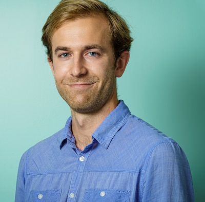 Headshot of a blond man wearing a blue button-down shirt. He stands against an aqua background.