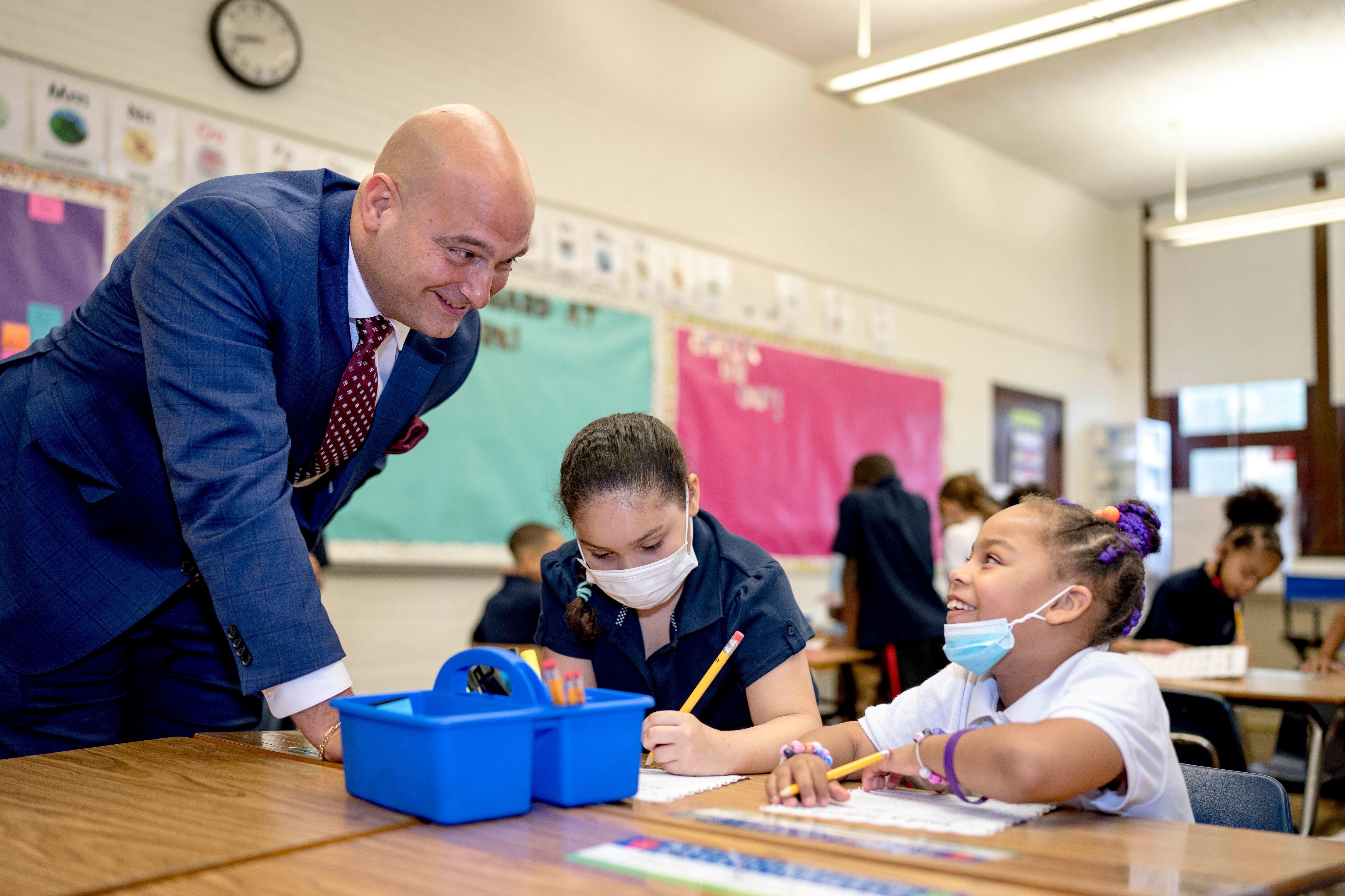 Detroit Superintendent Nikolai stands above two students working at a desk, talking with one of them as both students work on an assignment.