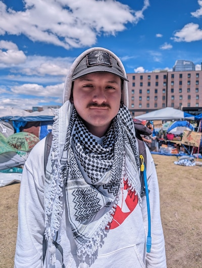 A student wearing a keffiyeh and a ball cap and hoodie faces the camera for a portrait with blue sky and clouds and buildings in the background.