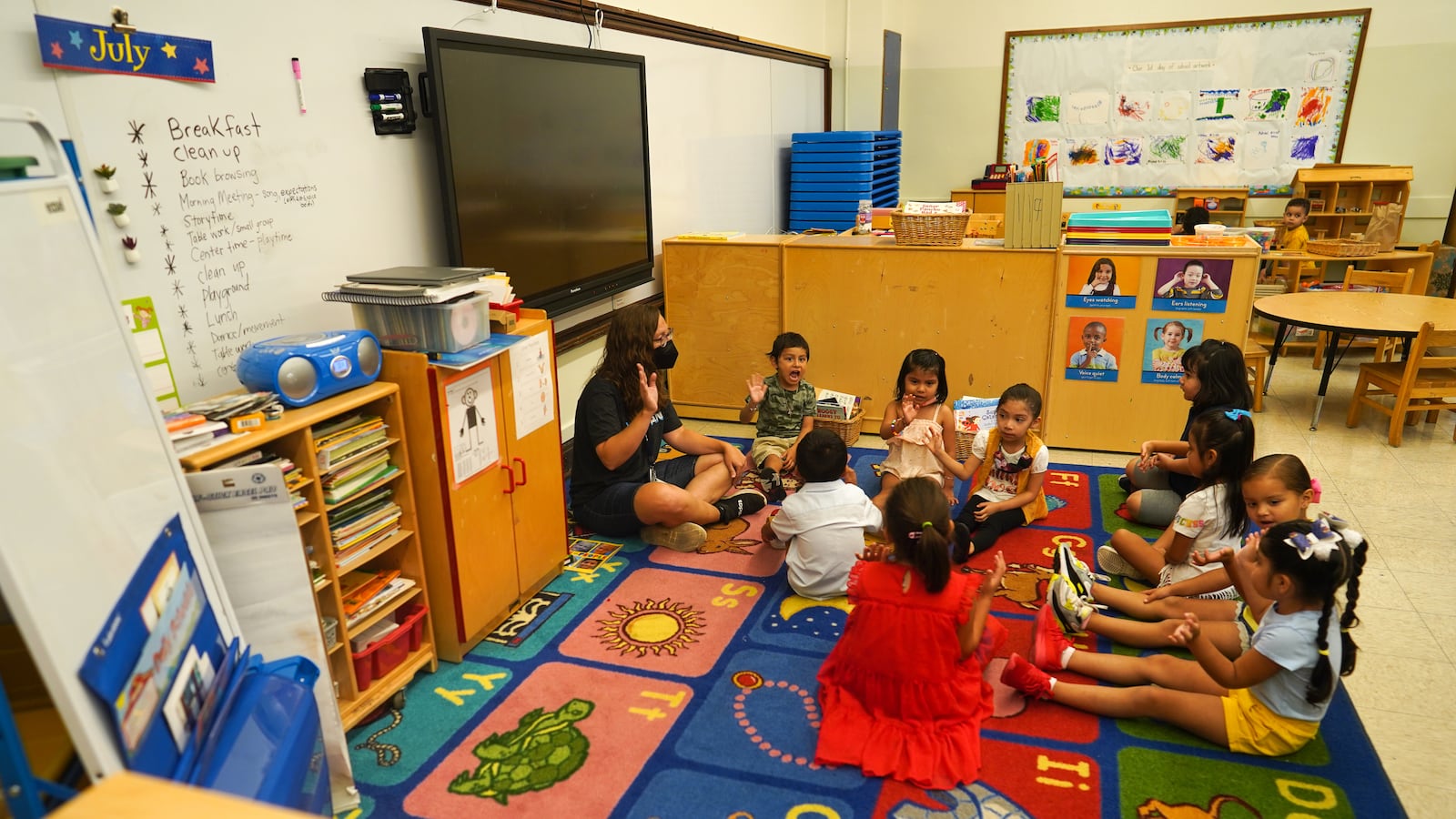 A group of young students and one teacher sit on a colorful rug in a classroom.