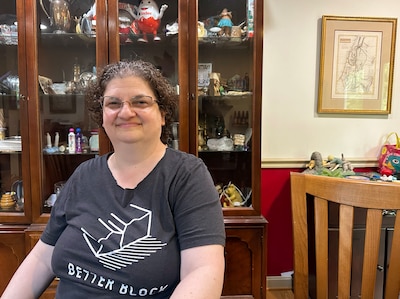 A woman with short dark hair and wearing a grey shirt with white writing poses for a photograph in front of a curio cabinet and a map of Palestine on the wall in the background.