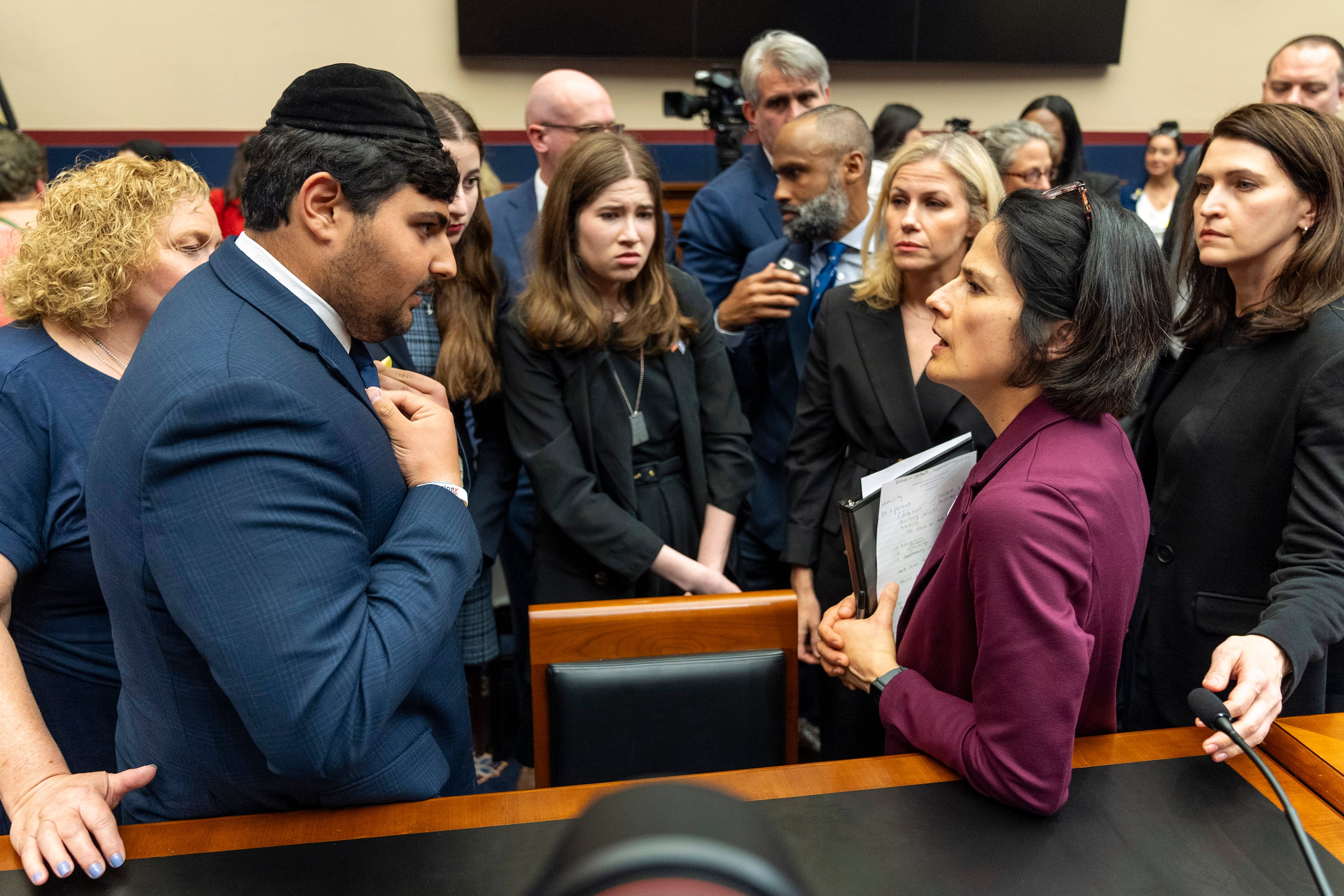 A teenage boy in a blue suit on the left faces a woman in a purple suit with a group of people in the background.