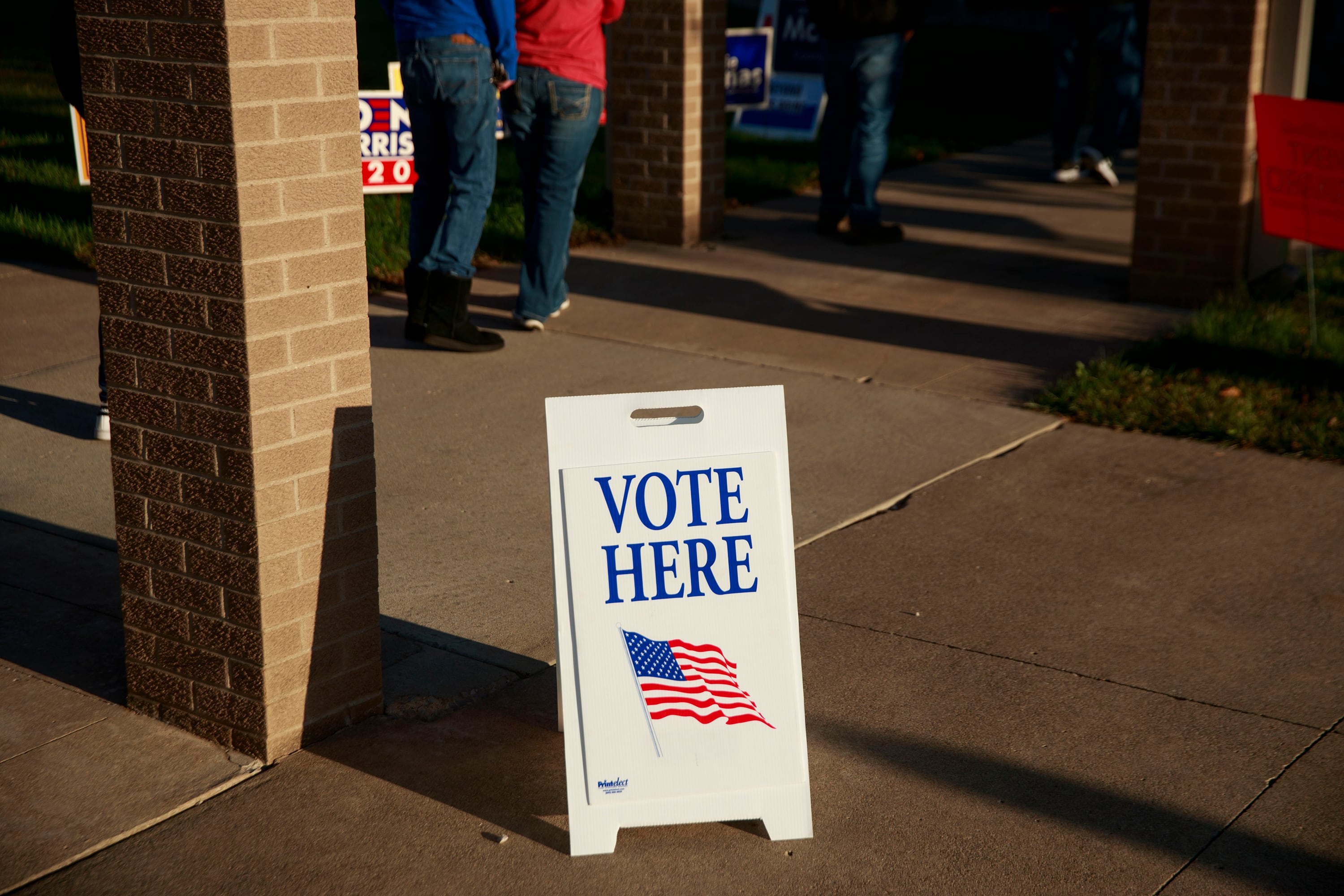 A sign that reads "Vote Here" with an American flag on it sits on a concrete sidewalk next to a building.