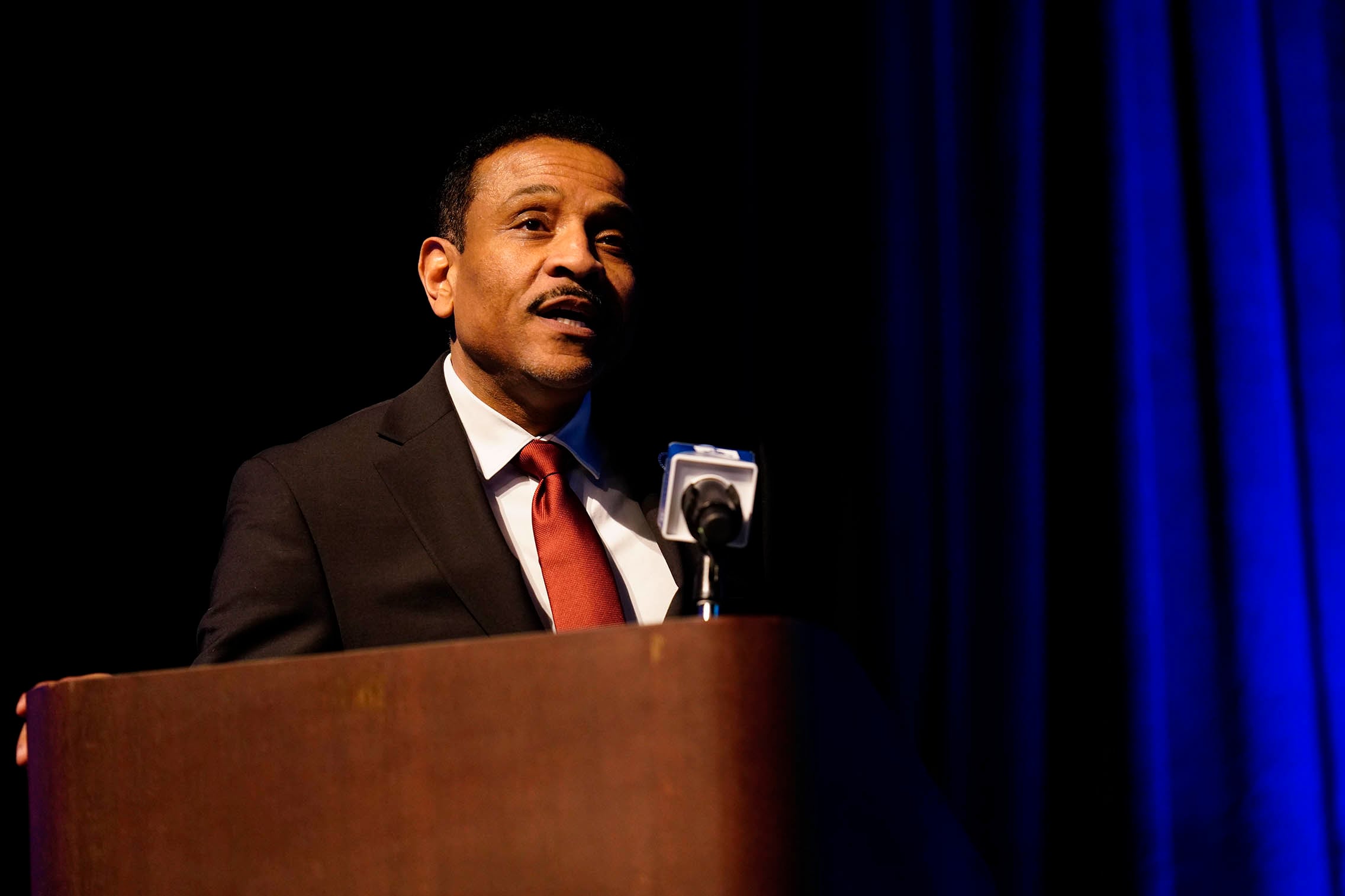 A man wearing a dark suit and a red tie stands at a wooden podium with a dark blue background.