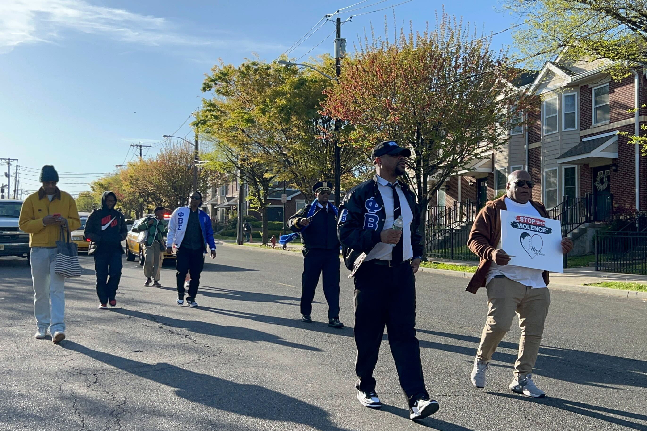 Several people walk down the middle of a street with a blue sky and a row of houses in the background.