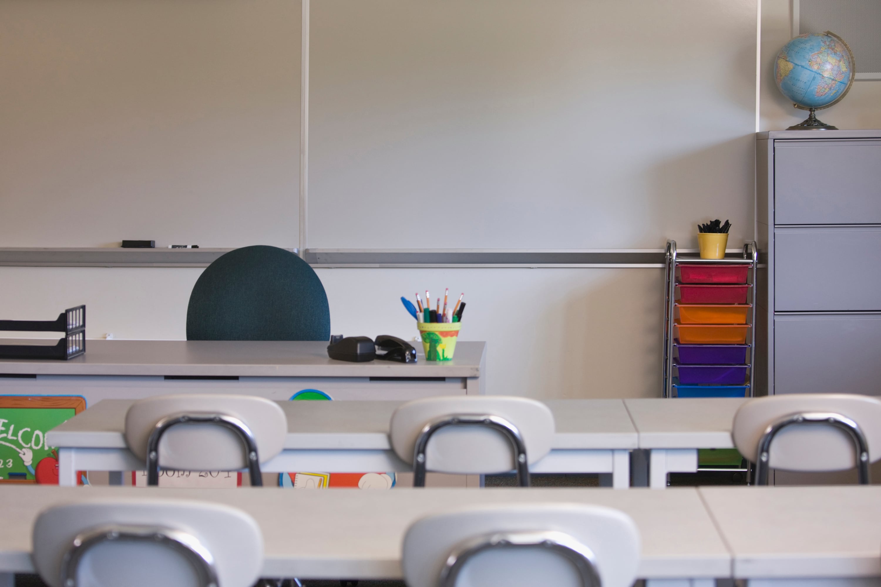 An empty class with a teacher's desk and chair in the background with two rows of desks and chairs in the foreground.