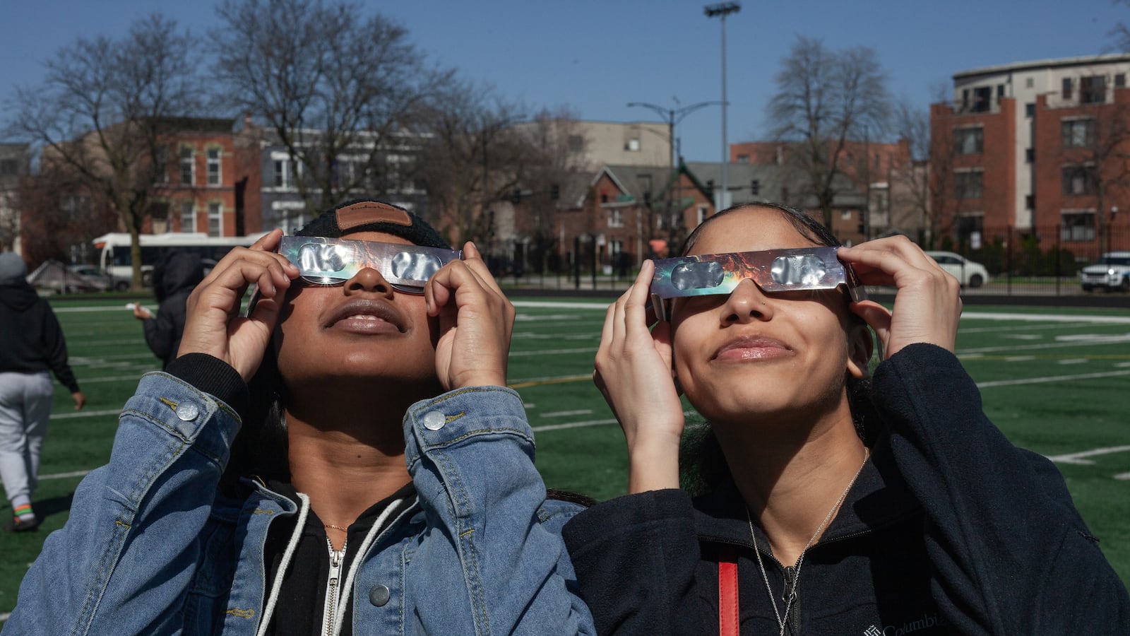 Two students holidng eclipse viewing glasses look up at the sky with a row of buidlings in the background.