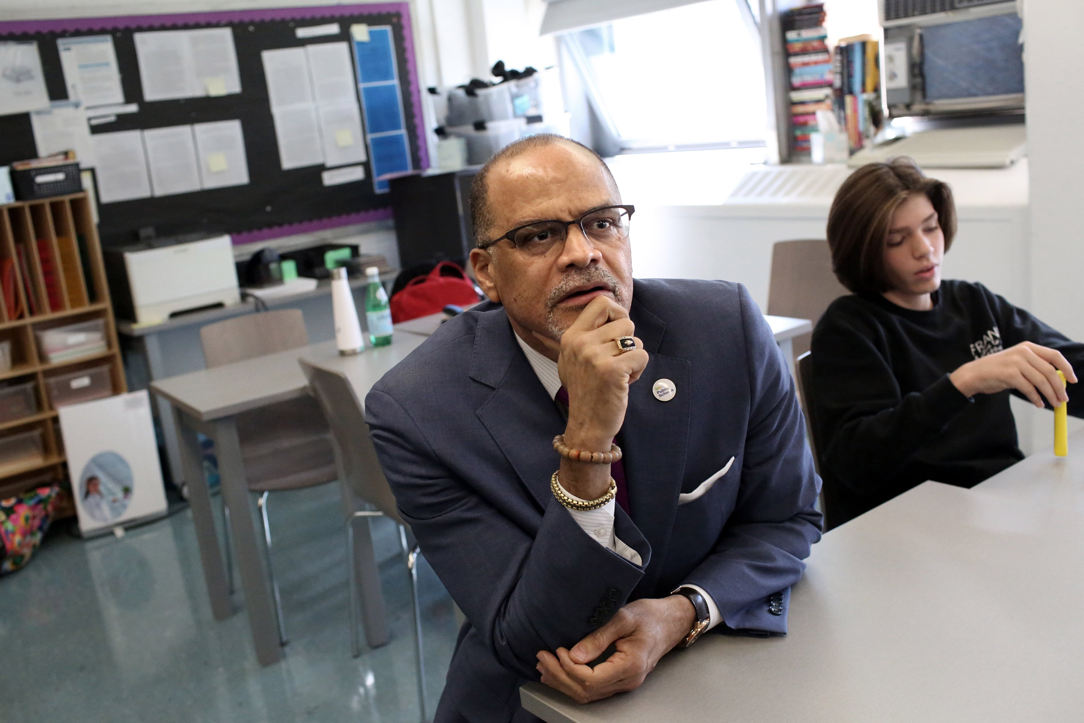 A man wearing a dark suit sits at a desk next to a student with a classroom scene in the background.