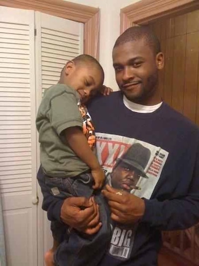 A man in a long sleeve blue t-shirt carries a toddler boy wearing a green t-shirt.