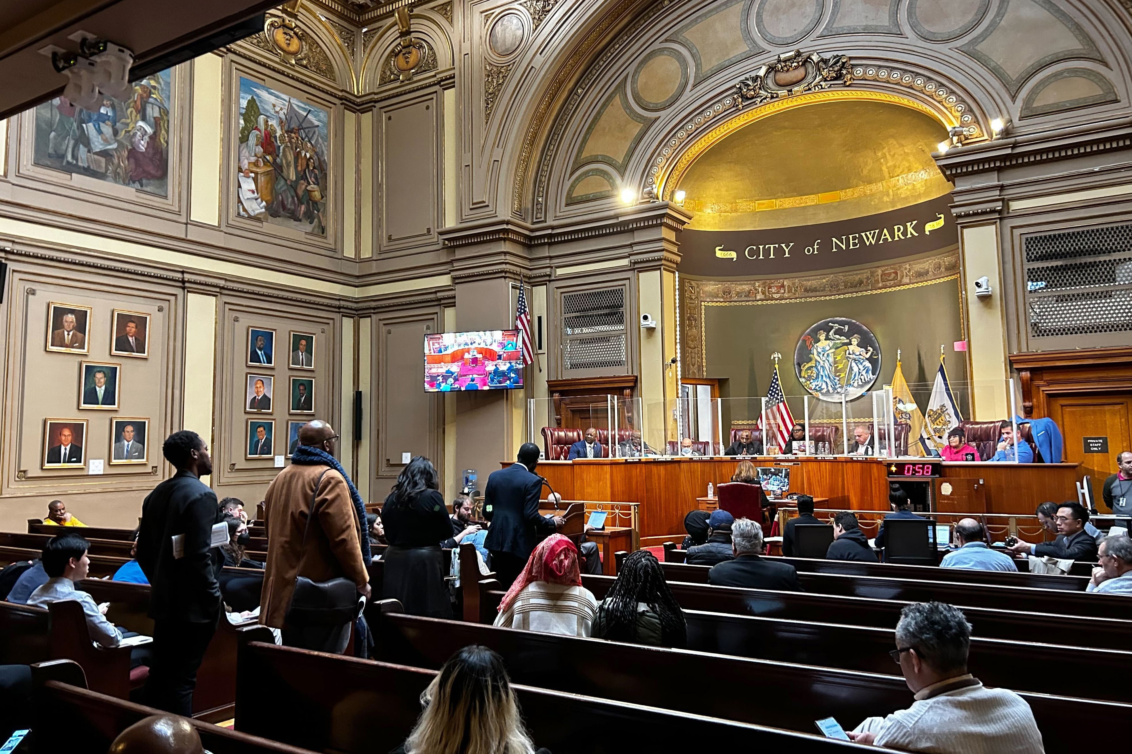 Several people stand in line while people sit in rows on each side of the line. A group of people sit at a wooden bench with the words "City of Newark" above them.