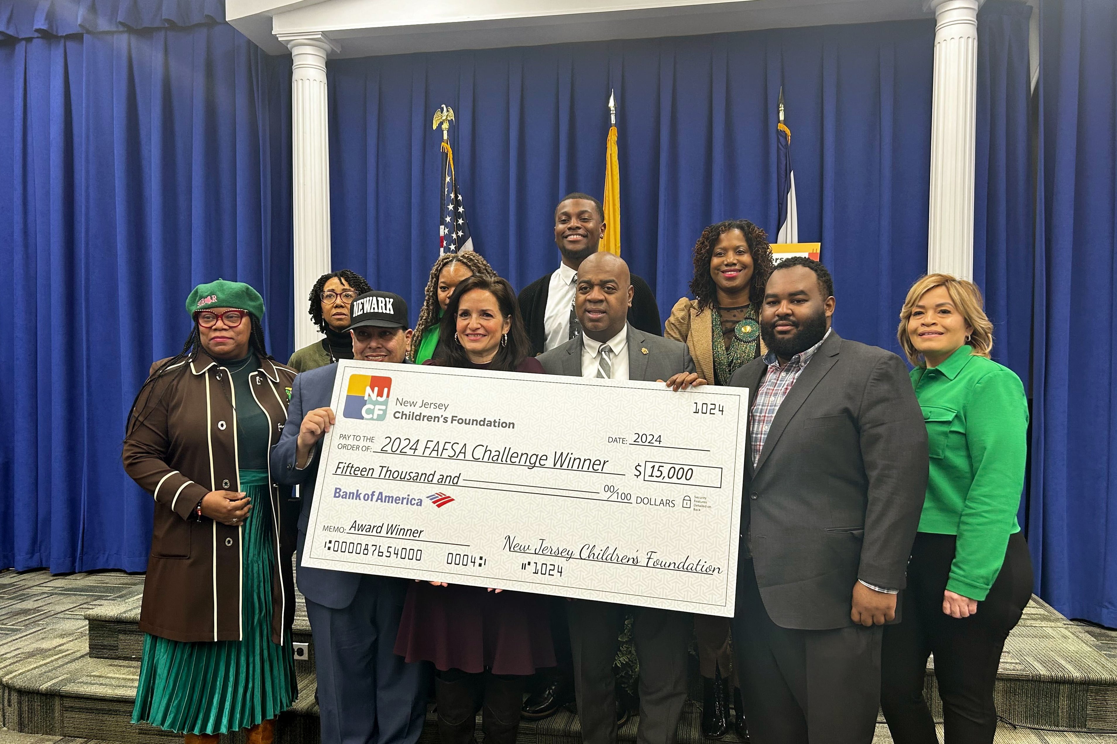 A group of people wearing suits and dresses stand together holding a giant check while they pose for a photograph. There are three flags and a dark blue curtain in the background.