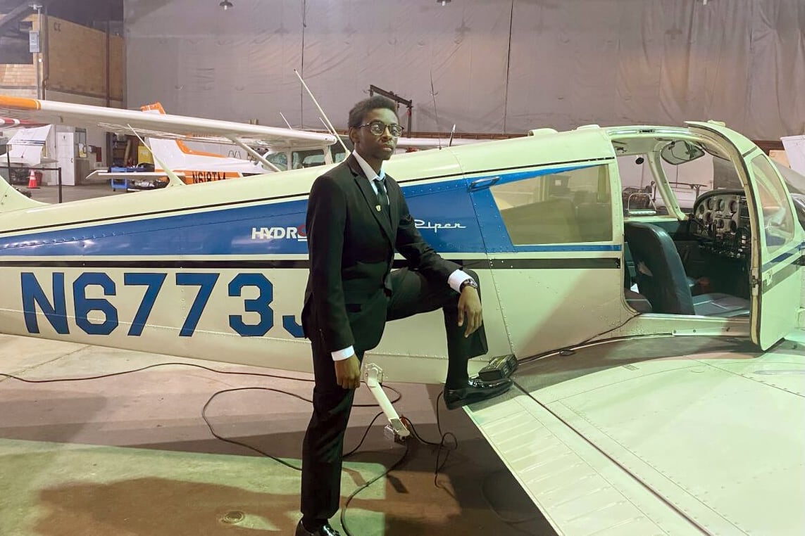 A high school student wearing a dark suit poses by the wing of a plane with a warehouse in the background.