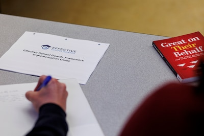 A hand is seen writing on a piece of paper with a packet of paper and a book next to them on the table.