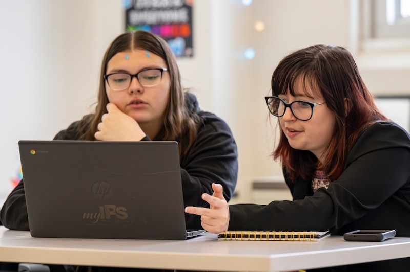 Two women sit looking at black laptop on a table.