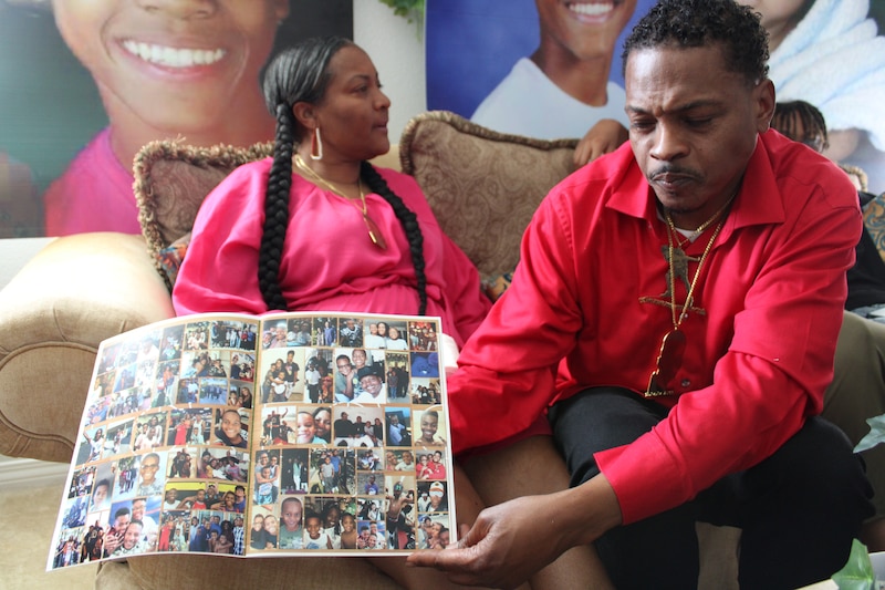 A man shows a photo album of a young boy, his son. On a couch behind him, the boy's mother looks on.