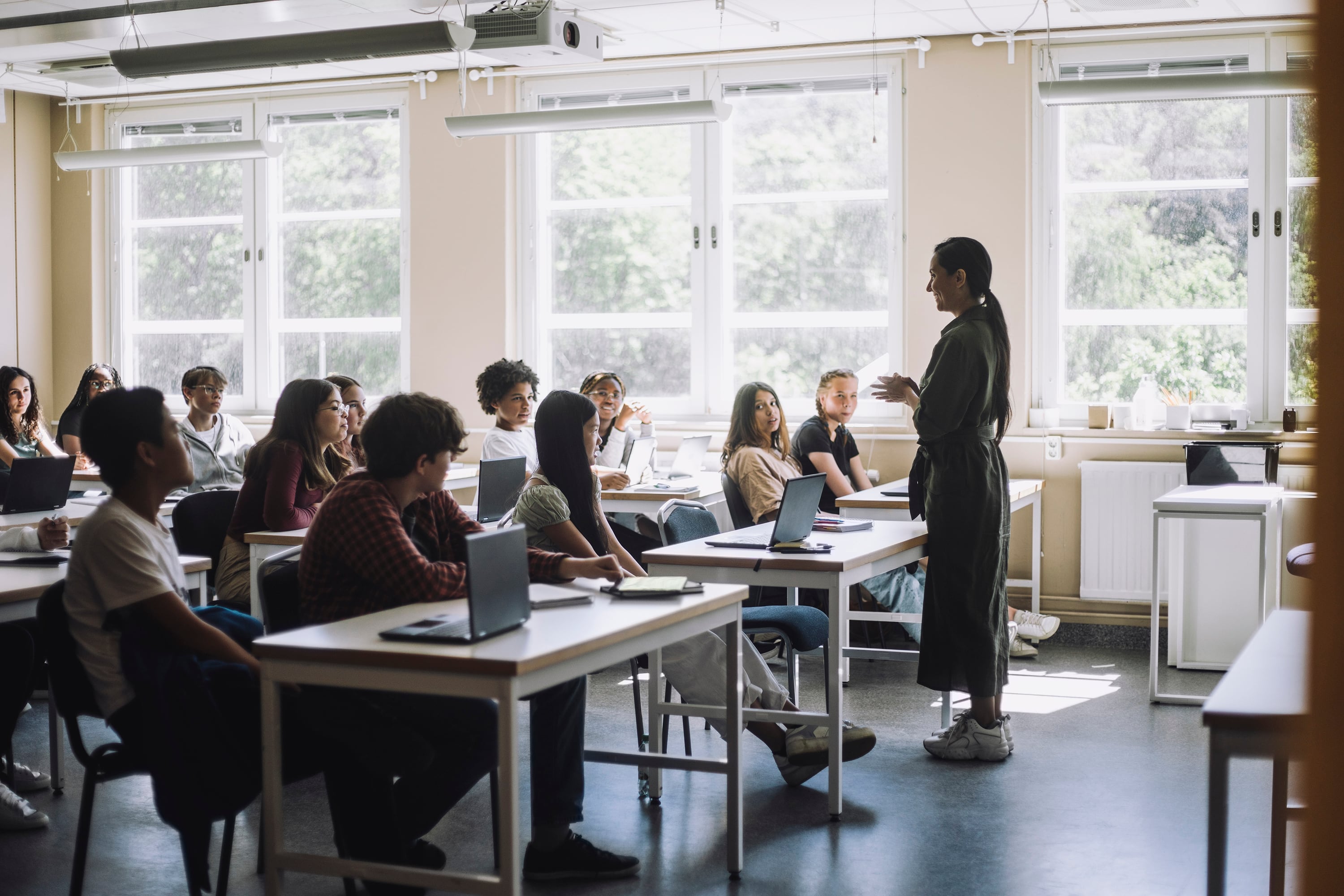 An adult stands at the right of the image facing a group of university students sitting at desks with large windows in the background.