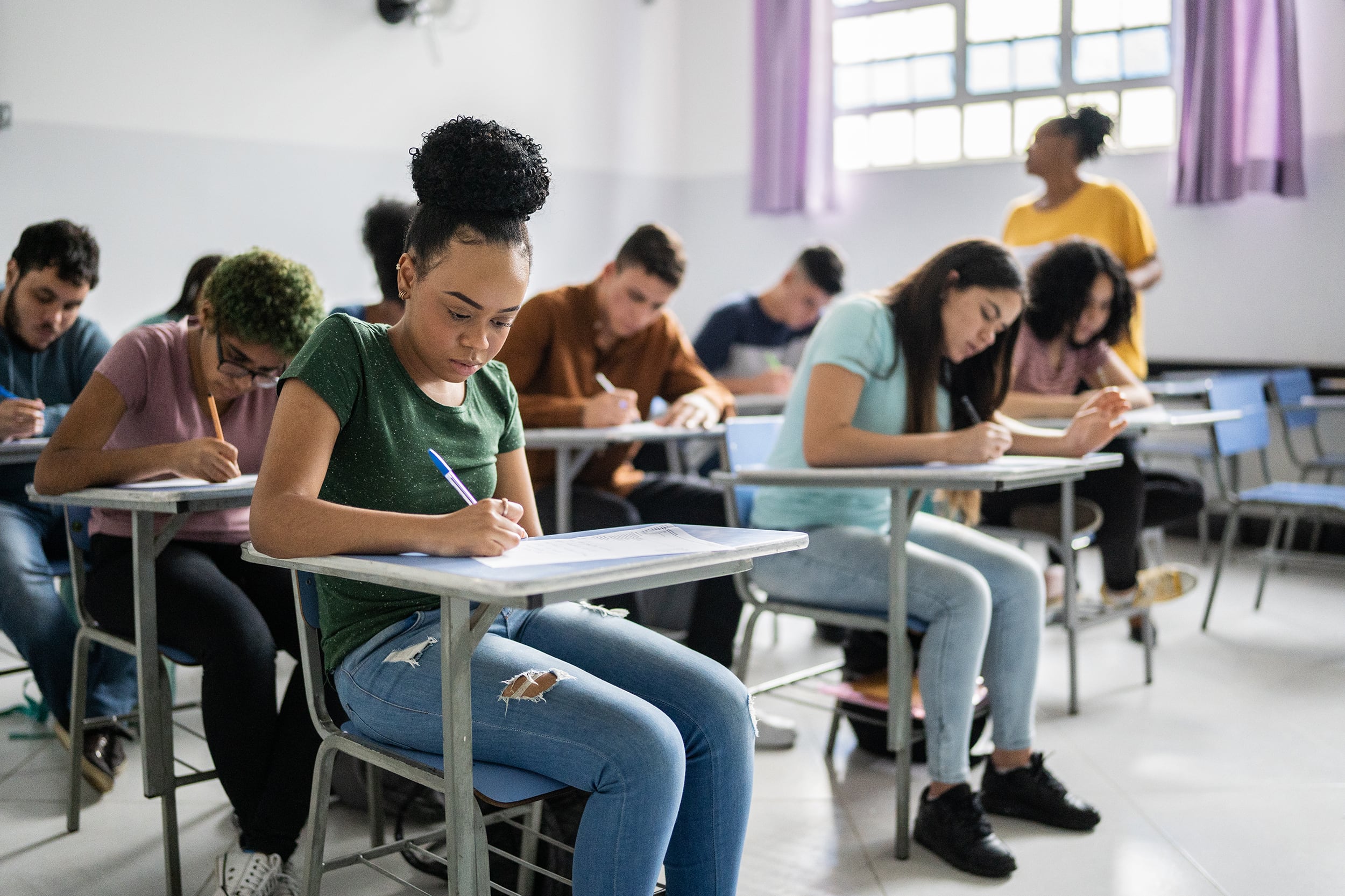 Several students write at desks in a classroom while a teacher walks by.