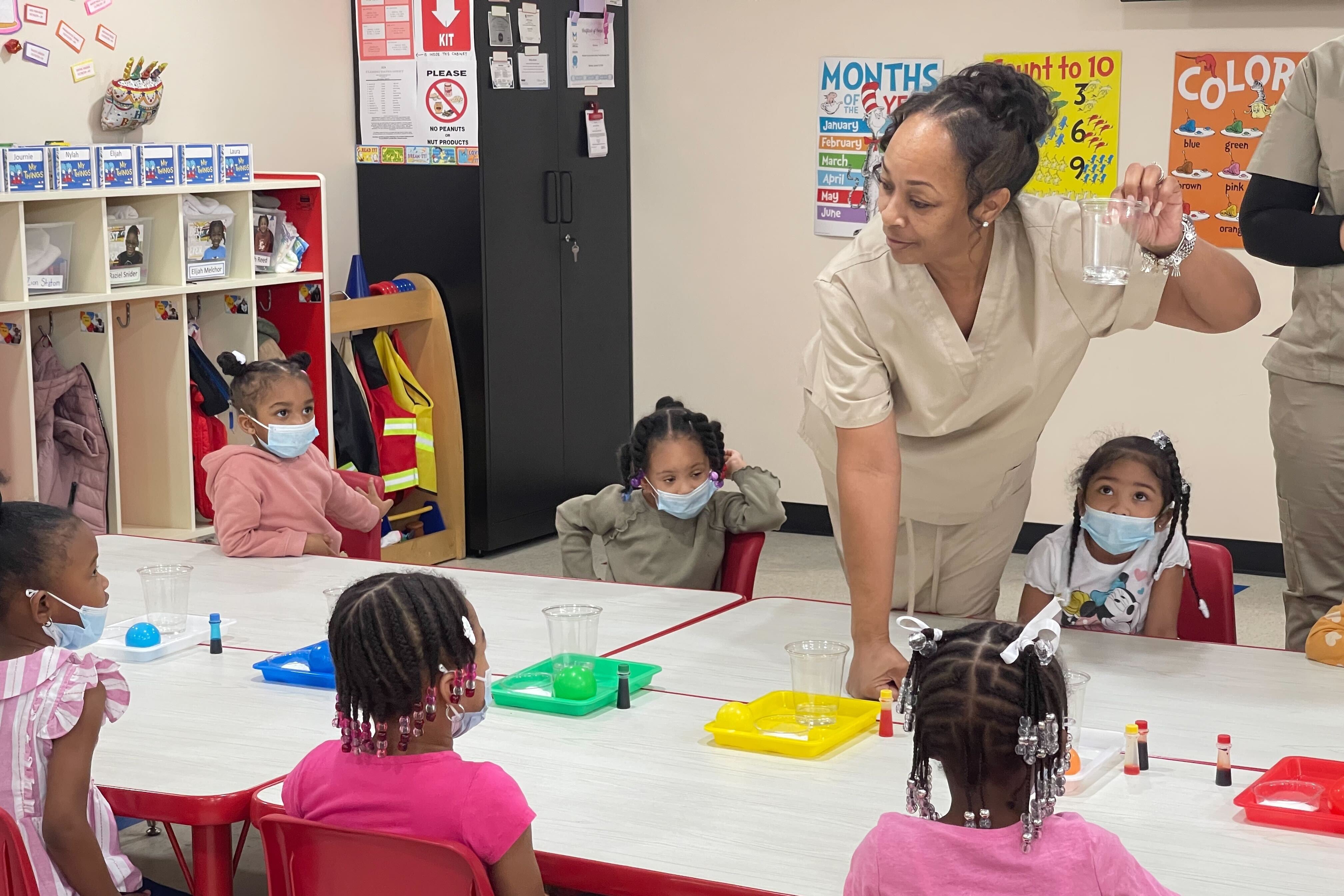 Students in a prekindergarten classroom in Detroit sit at a table and listen to their teacher who is holding a cup.