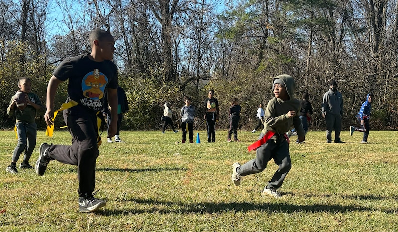 Two young boys play flag football on a green grassy field with other young boys in the background.