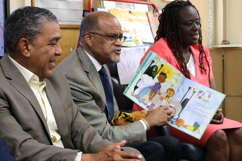 Three adults wearing business clothes sit side by side in a classroom while the middle person is holding a book.