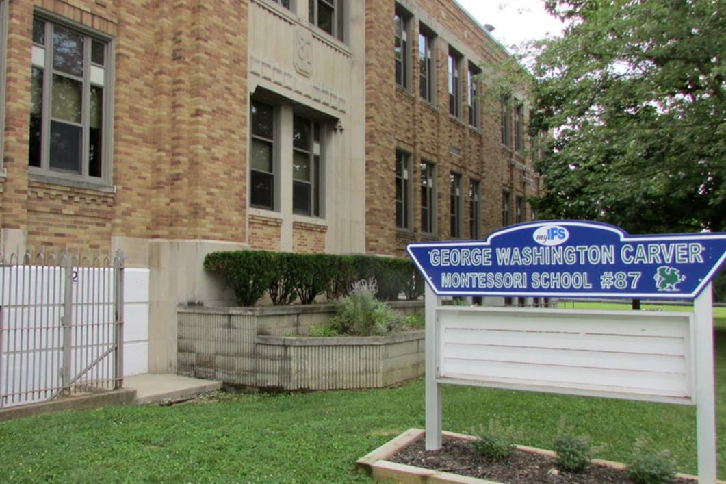 A  white and blue school sign stands on a green lawn in front of a large, tan, brick building.