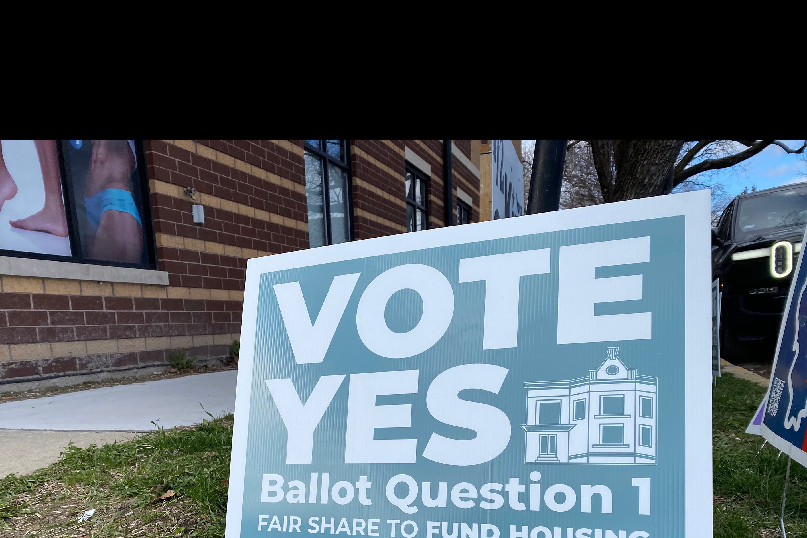 A blue and white sign sits in a grassy lawn outside of a red brick building. Some of the words on the sign read "Vote Yes Ballot Question 1."