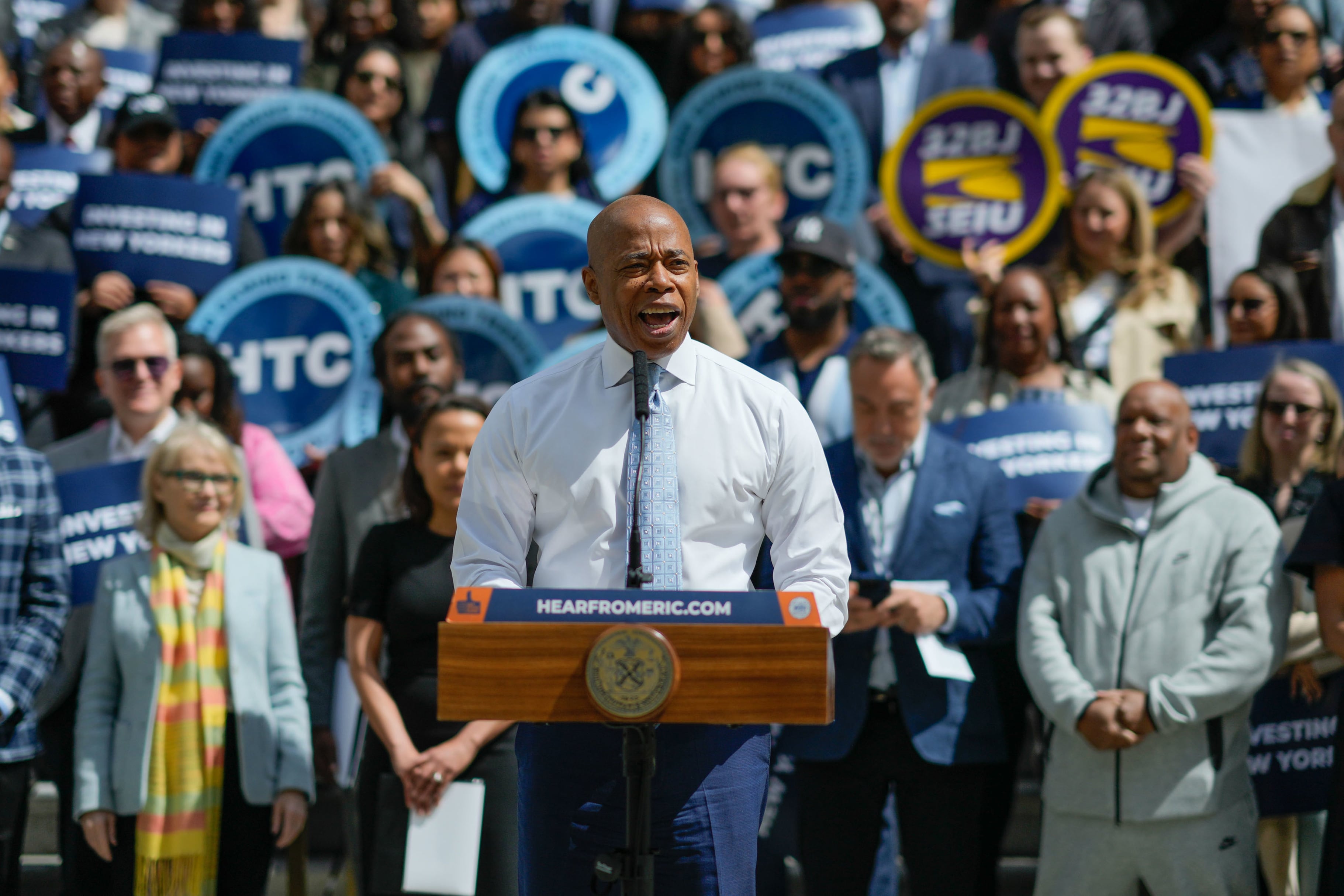 A man in a white dress shirt stands at a podium with a crowd behind him.