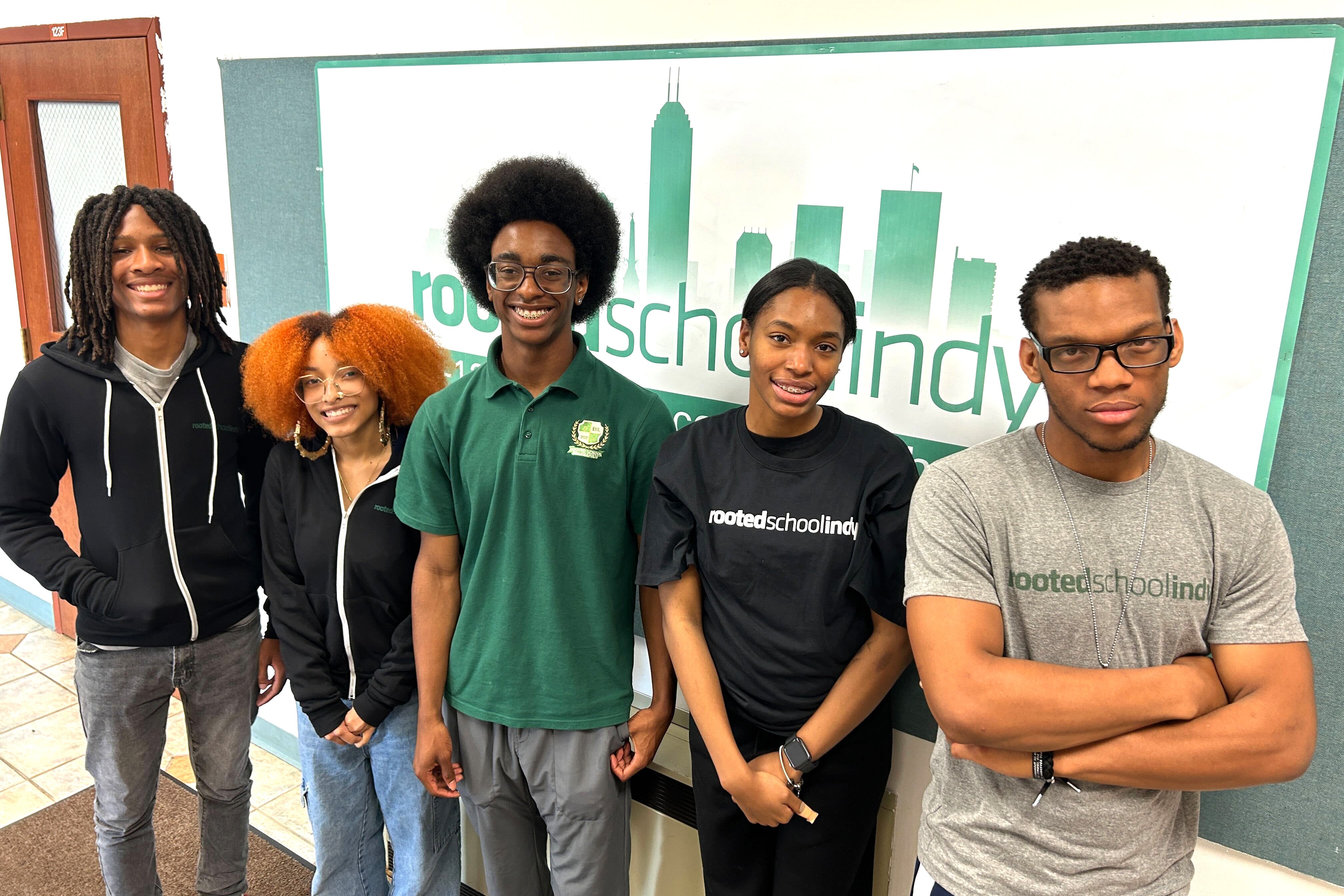 A male student in a black sweat jacket, a female student in a black sweat jacket, a male student in a green shirt, a female student in a black t-shirt and a male student in a gray t-shirt stand in front of a sign.