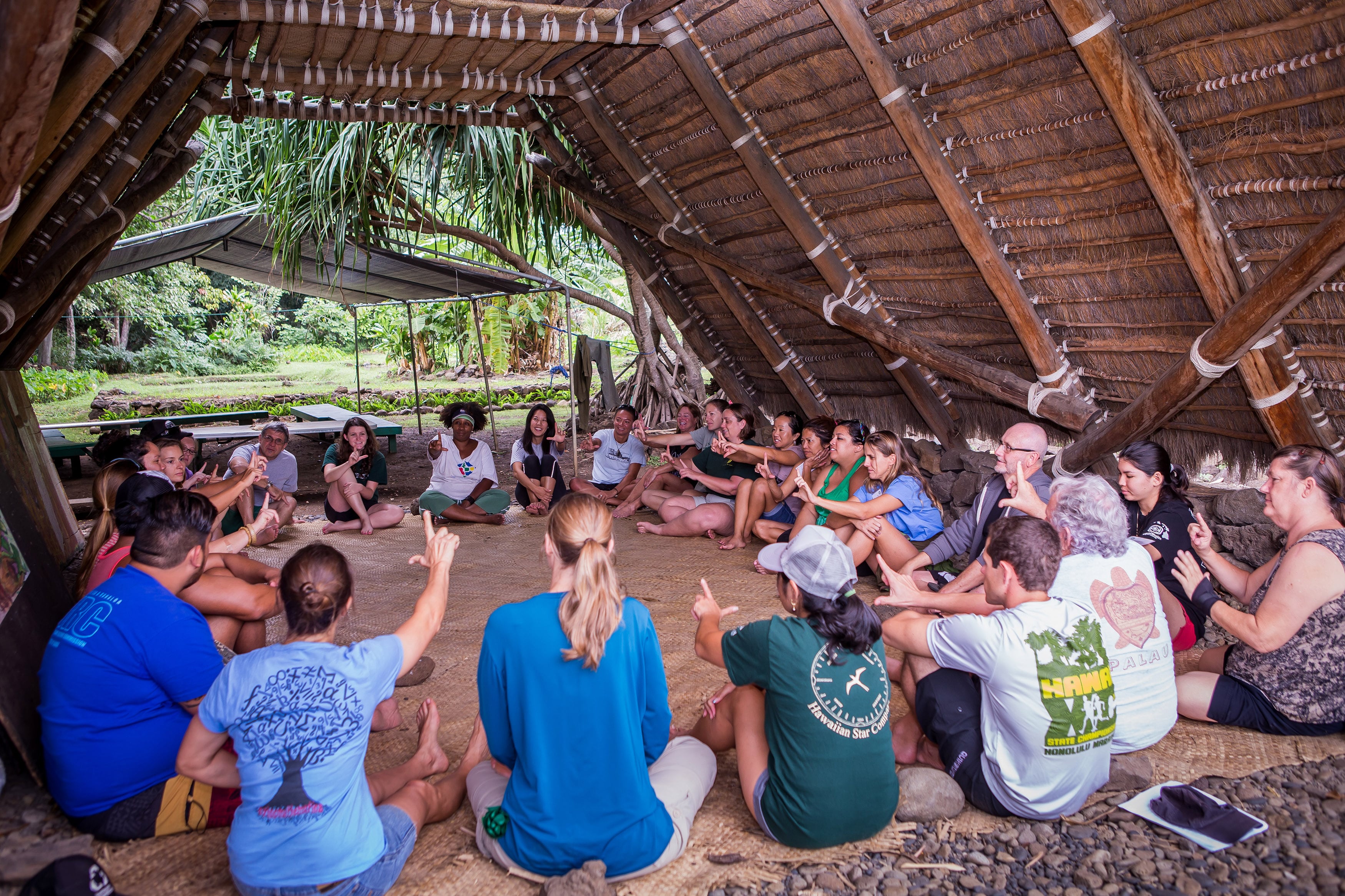 A group of people sit on the ground in a circle under a wooden structure.