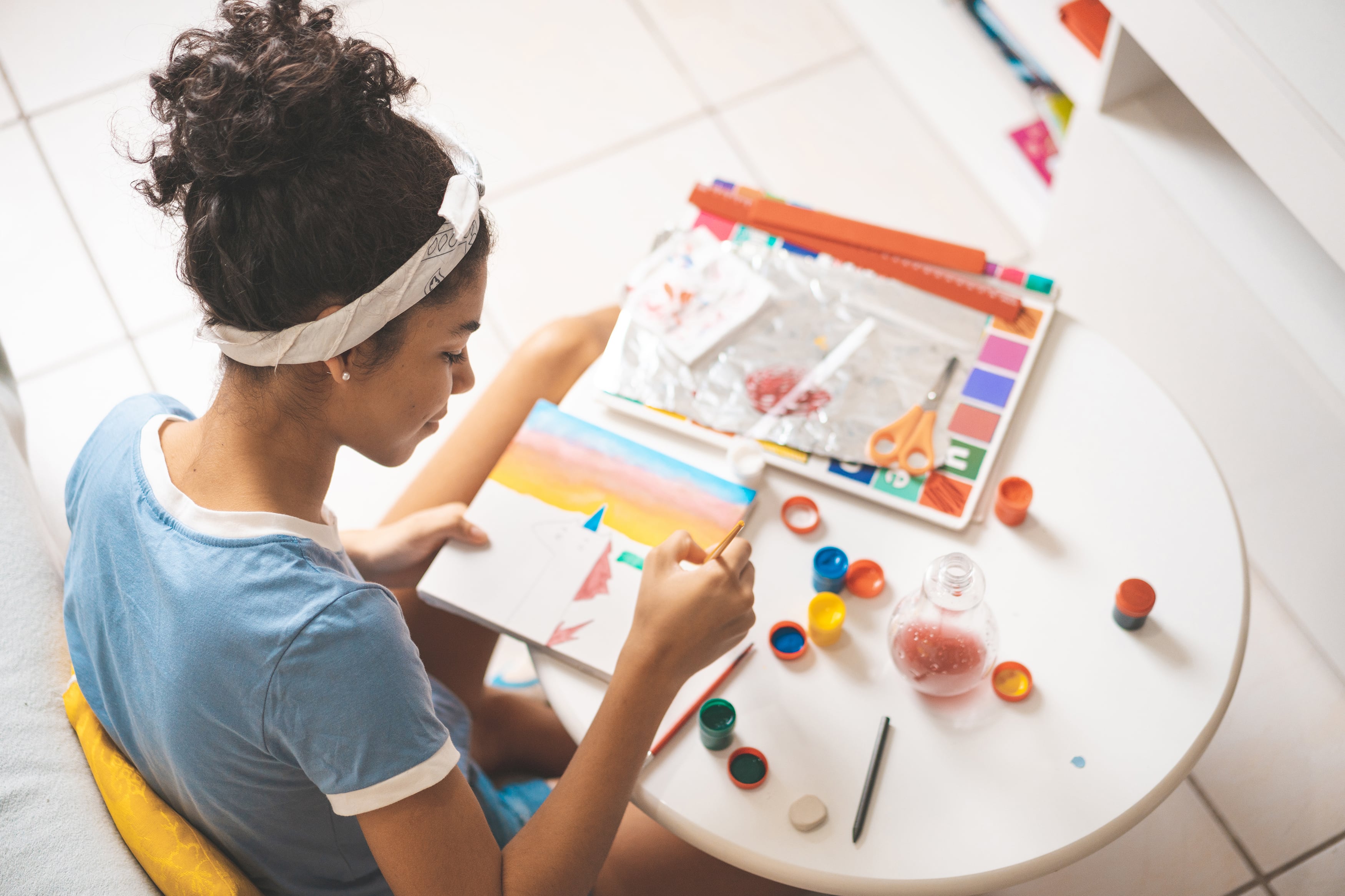 A bird's eye view of a high school teen with curly, dark hair in a bun sits at a round white table painting.