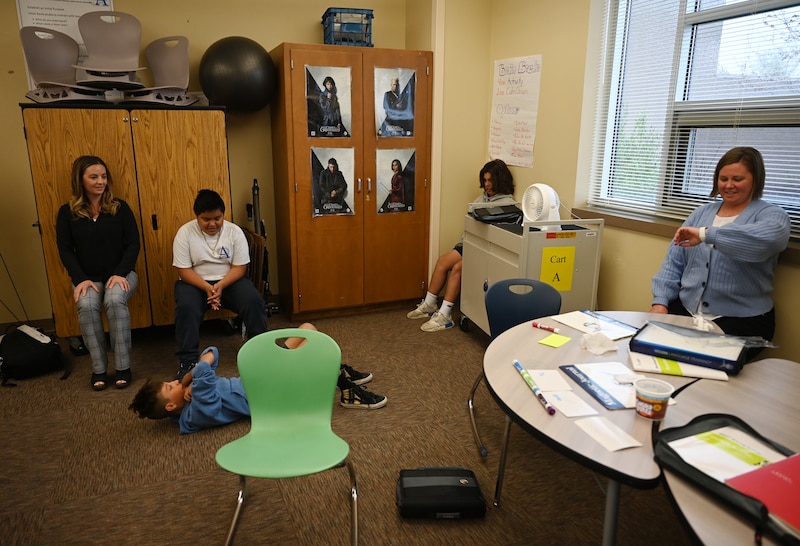 Three students and their two teachers take a break during a lesson in their classroom, as one student wearing a blue sweatshirt lies on the floor.