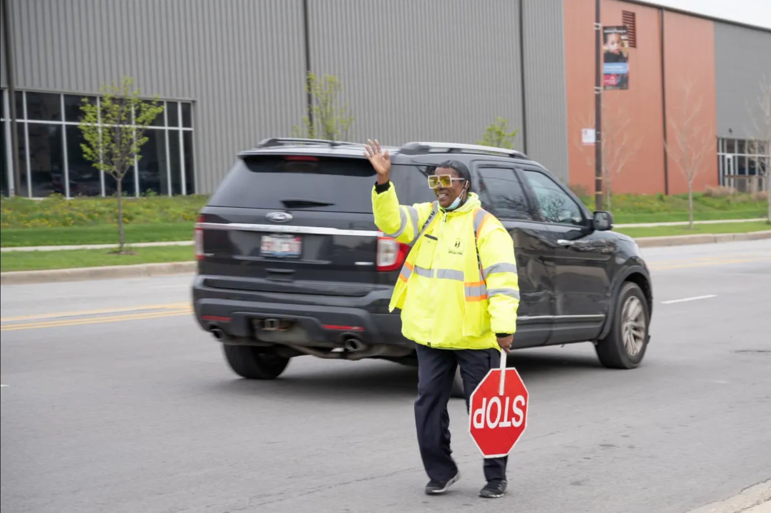 A person wearing a yellow safety jacket and hat and holding a stop sign waves and stands in the middle of the street.