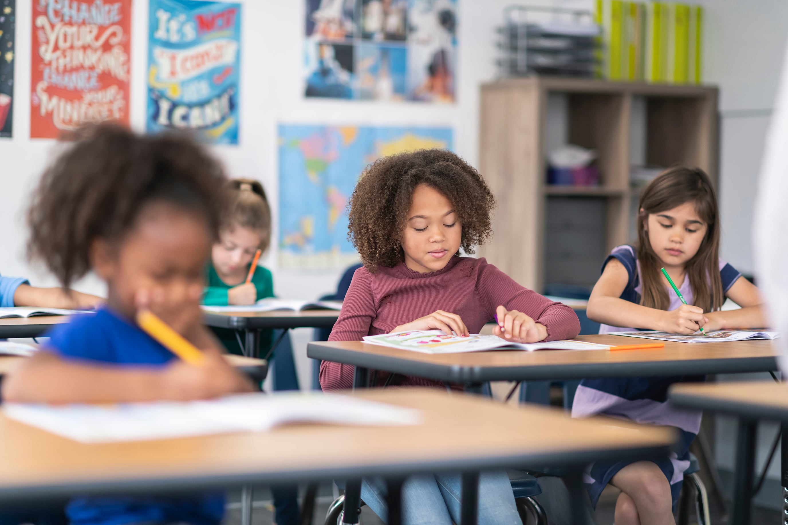 Three young students work at their desks in a classroom with posters on the back wall.