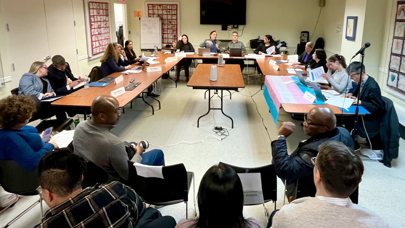 A group of people sit at tables in a rectangle in a room.