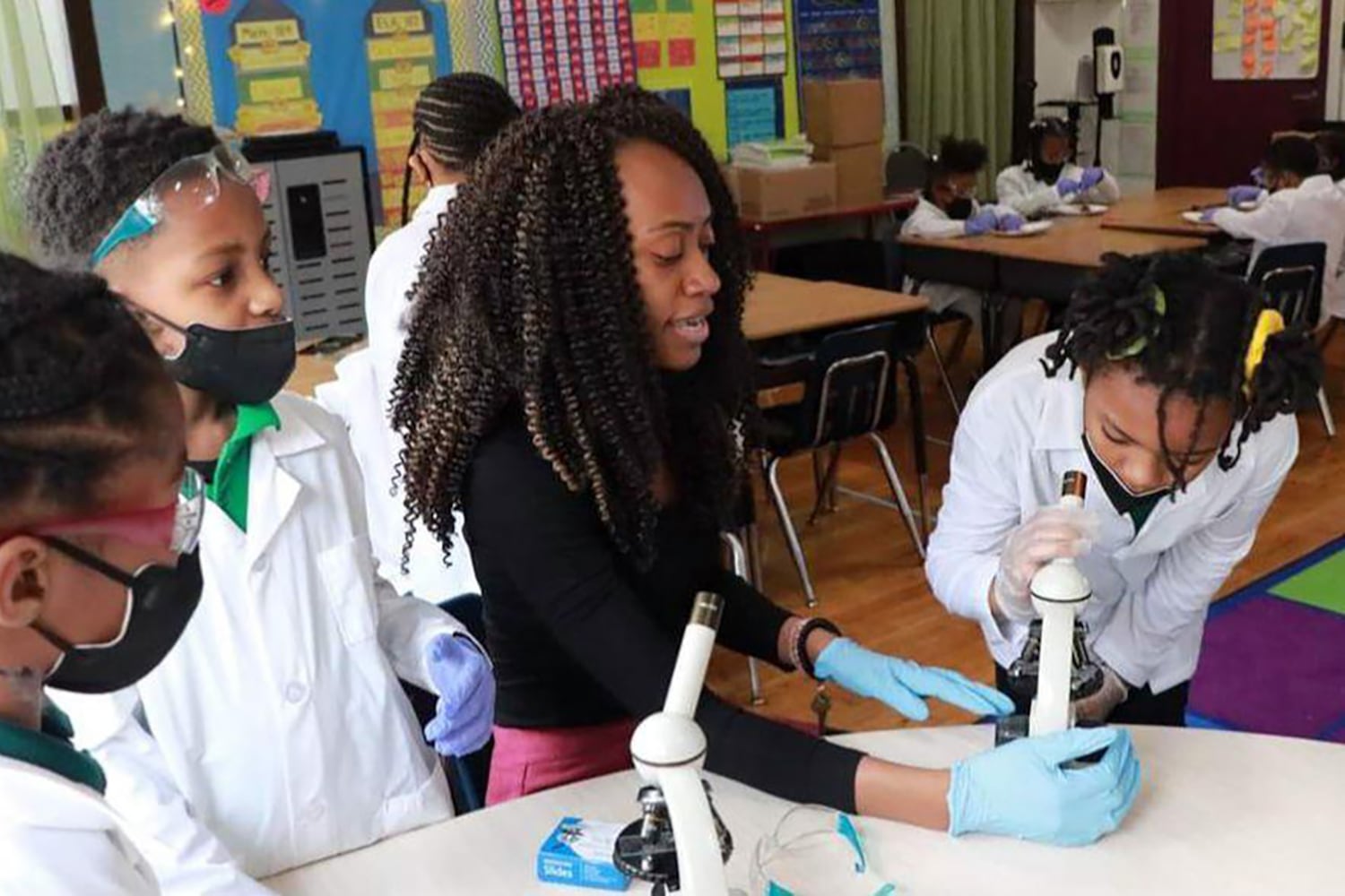 An adult with long, dark, curly hair and wearing a black blouse, sits at a table with three young students wearing lab coats and all working together in a classroom.