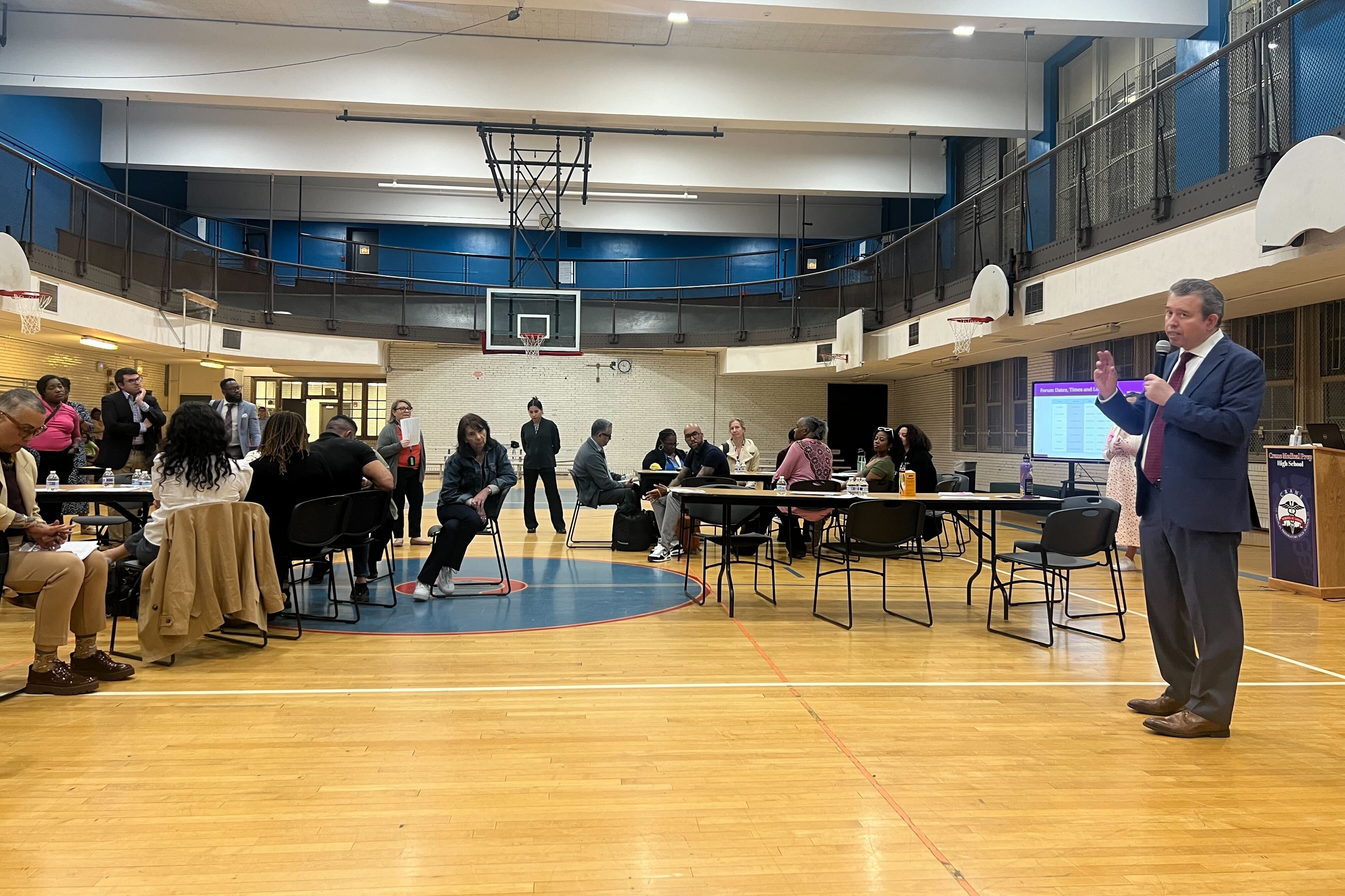 A man in a suit is standing up in a school gym speaking to people seated at tables.