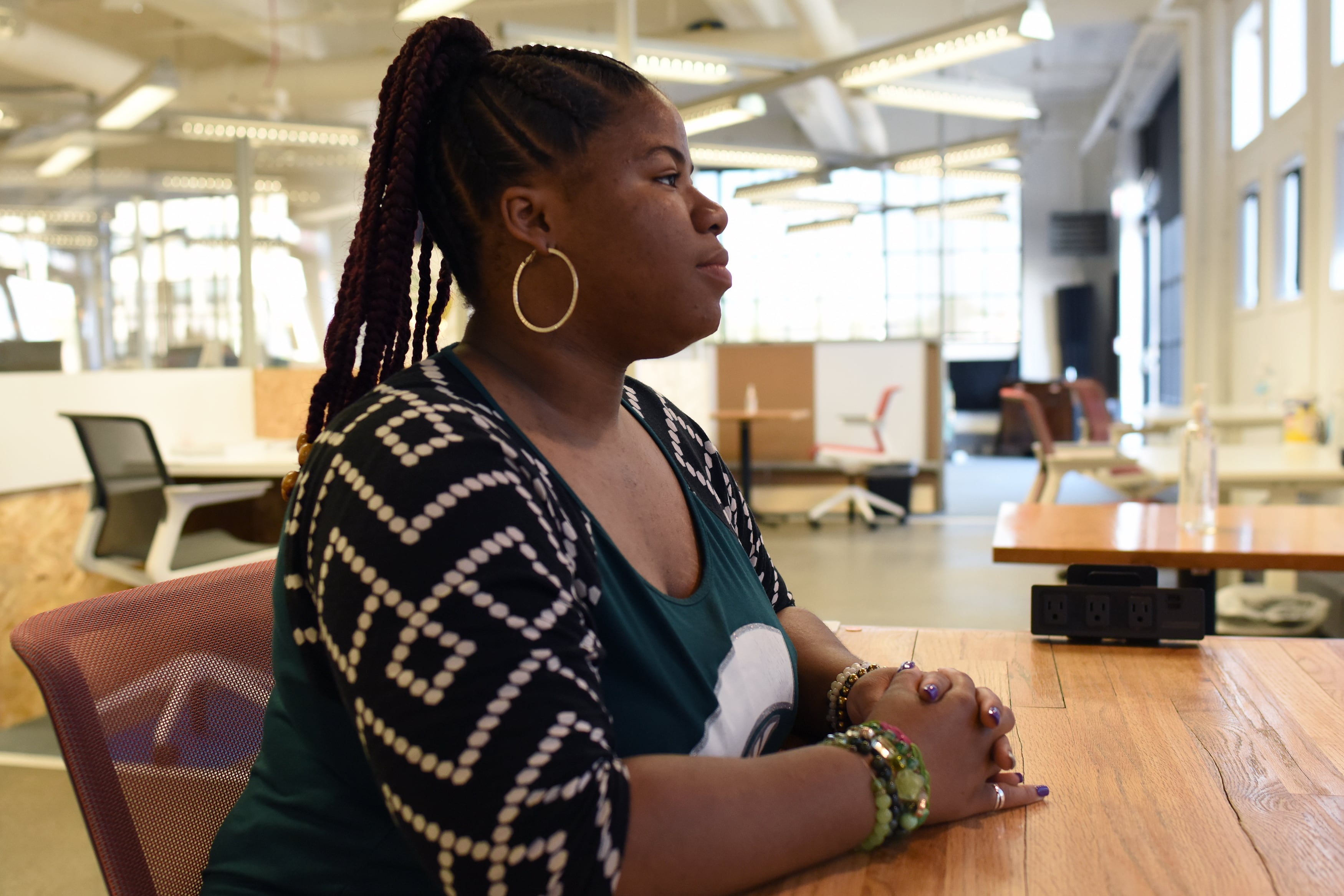 A high school senior girl with long dark braids pulled up into a pony tail, wearing a black, white and green shirt poses for a portrait at a wooden table in side of a school.