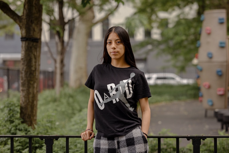 A high school student with long dark hair and wearing a black t-shirt, poses for a portrait while leaning up against a black metal fence with green trees and a car in the background.