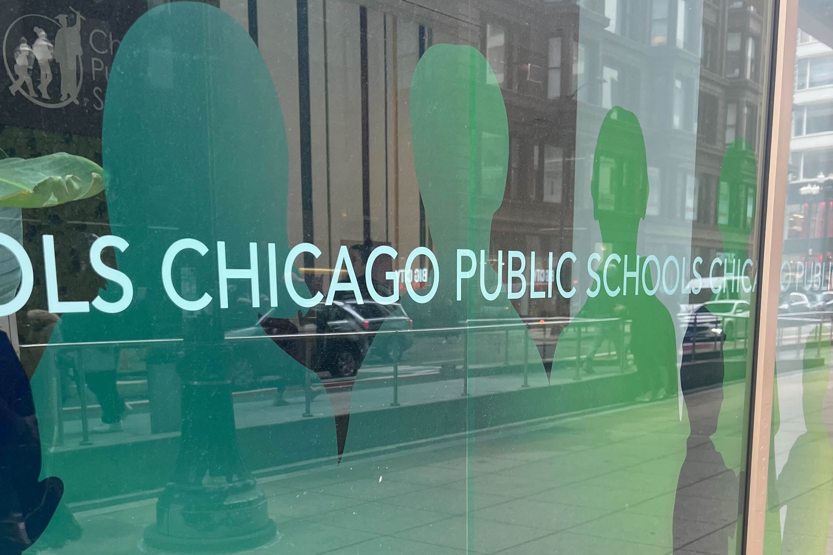 The Chicago Public Schools logo on the side of a glass wall with reflection of cars and people passing in the background.