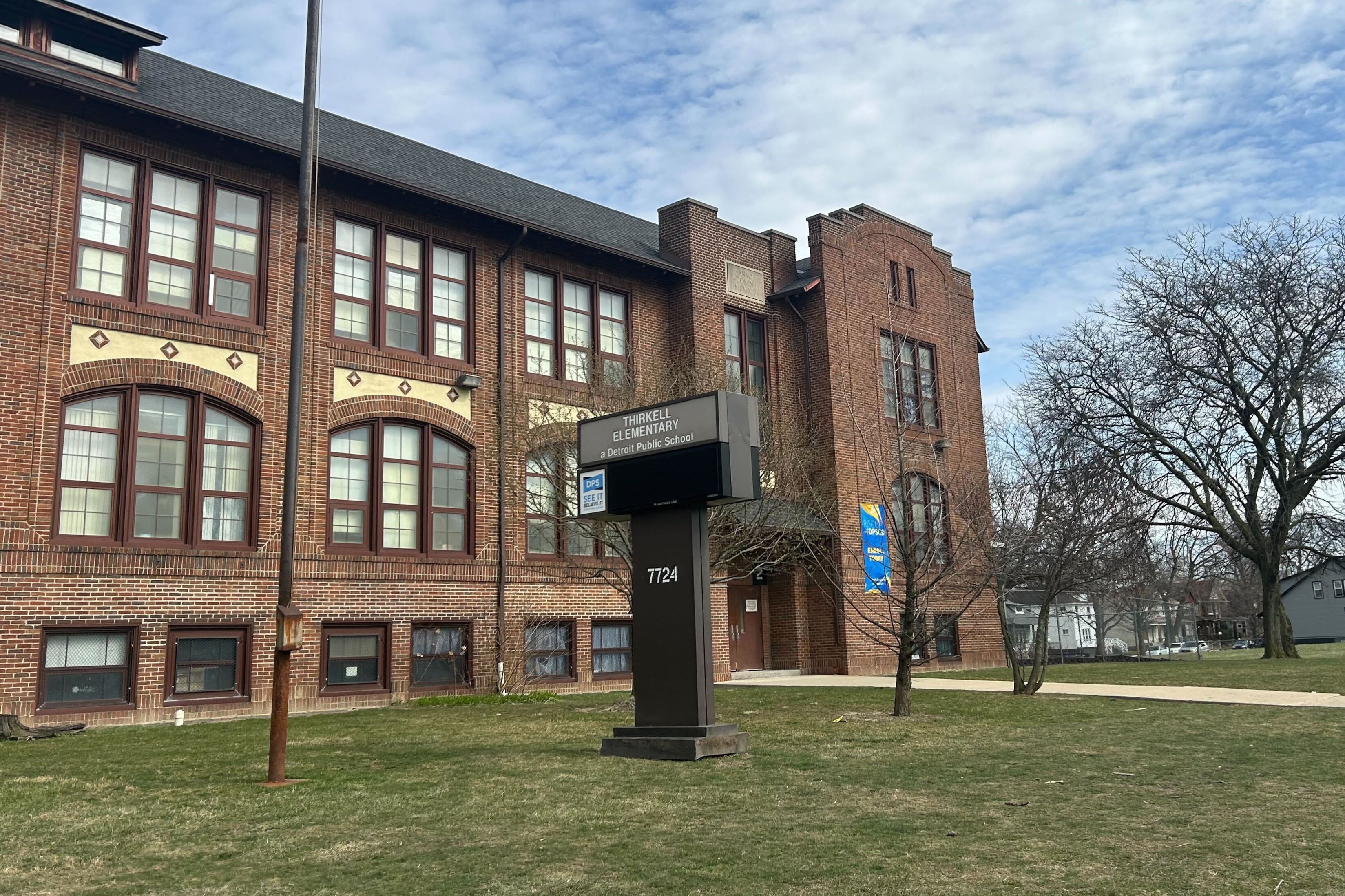 A large red brick building with a sign in a grassy field in the front and a blue and cloudy sky in the background.