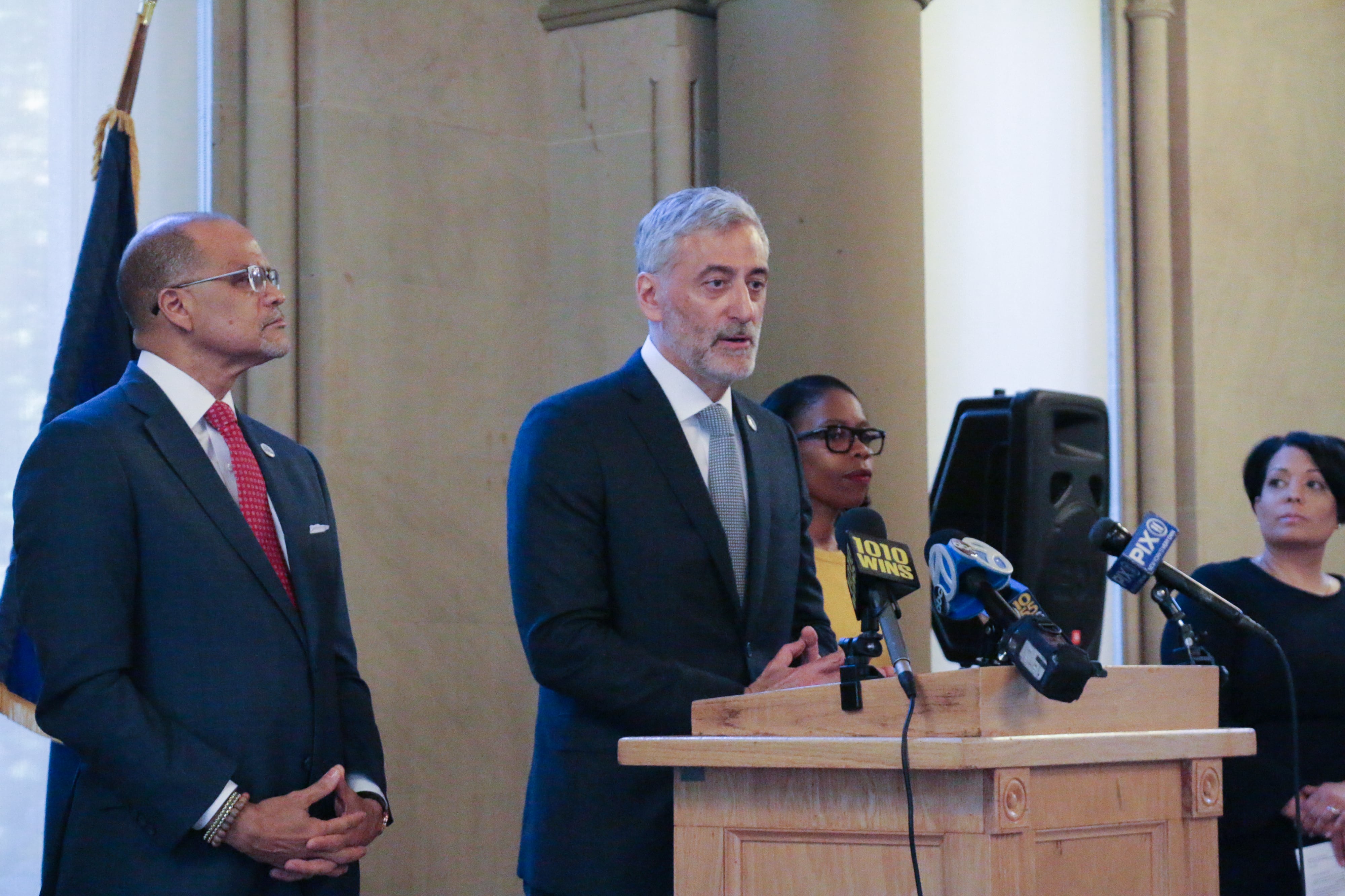 Four people in business clothes stand side by side with one person at a podium speaking.