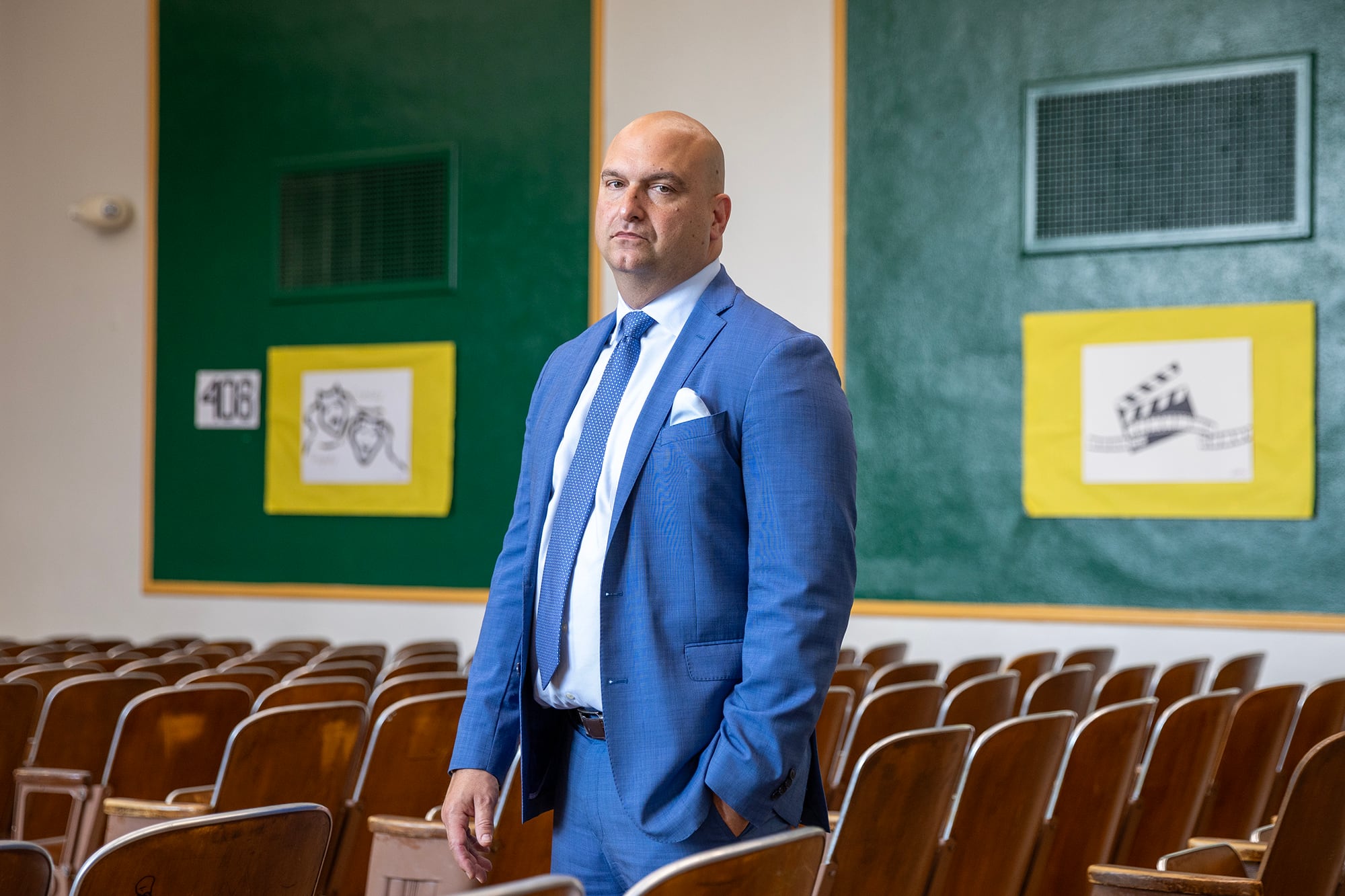 A man with no hair and wearing a dark suit poses for a portrait while standing in a row of chairs in a school auditorium.