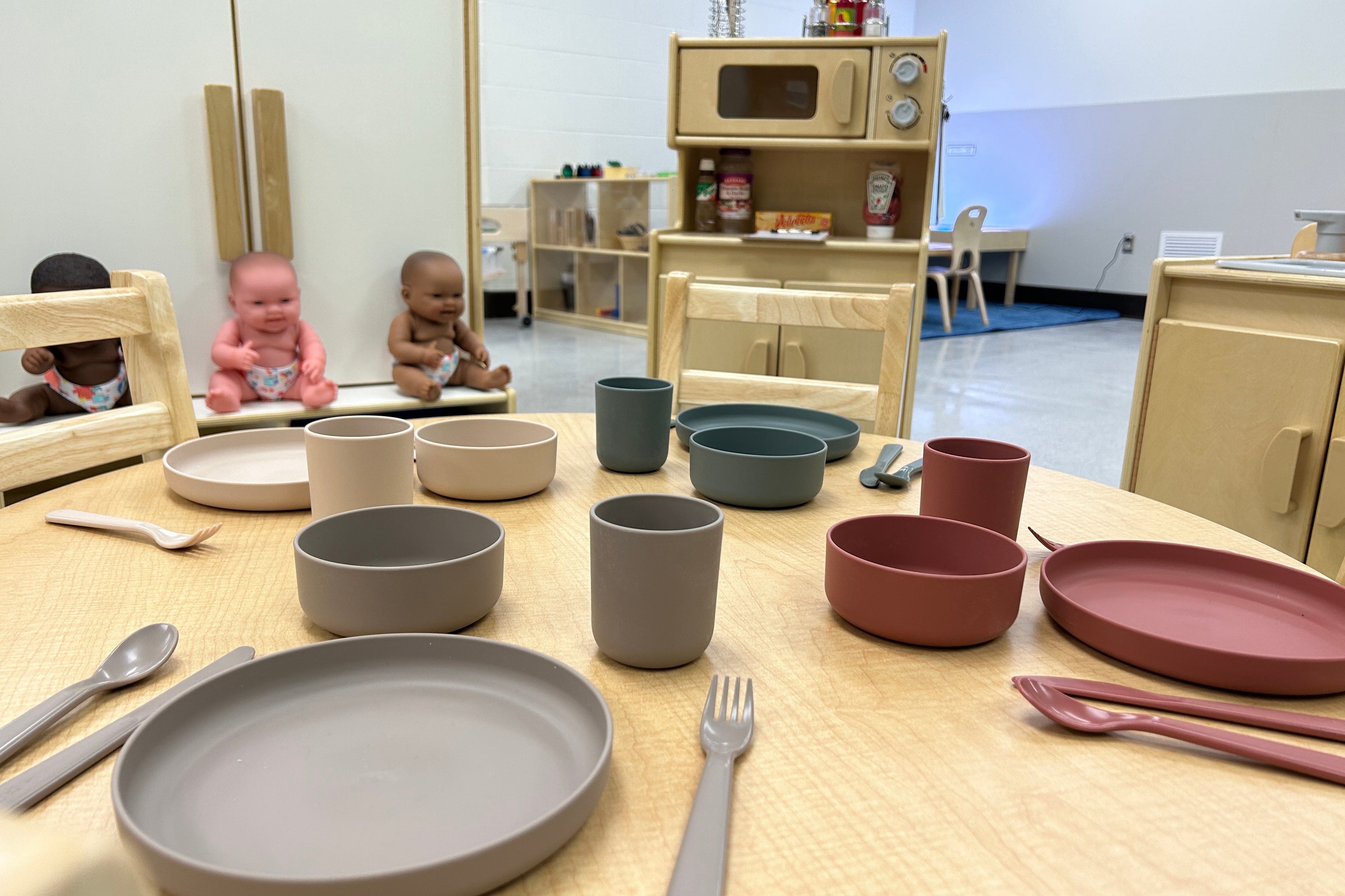 Silverware and plates sit on a children’s dining table in the foreground, with children’s kitchen playware in the background.