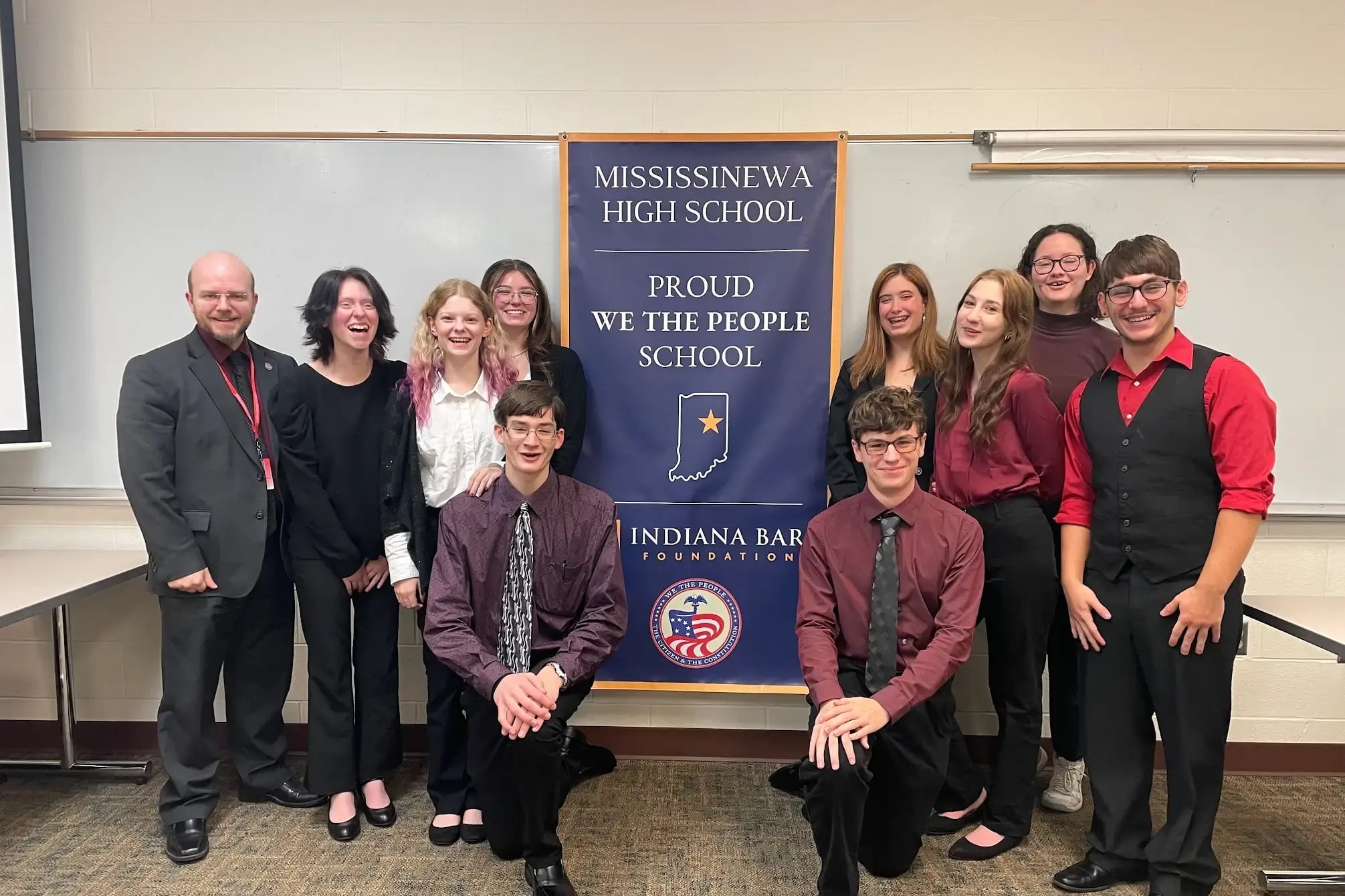 Teachers and students stand next to a sign that reads "Proud We The People School."